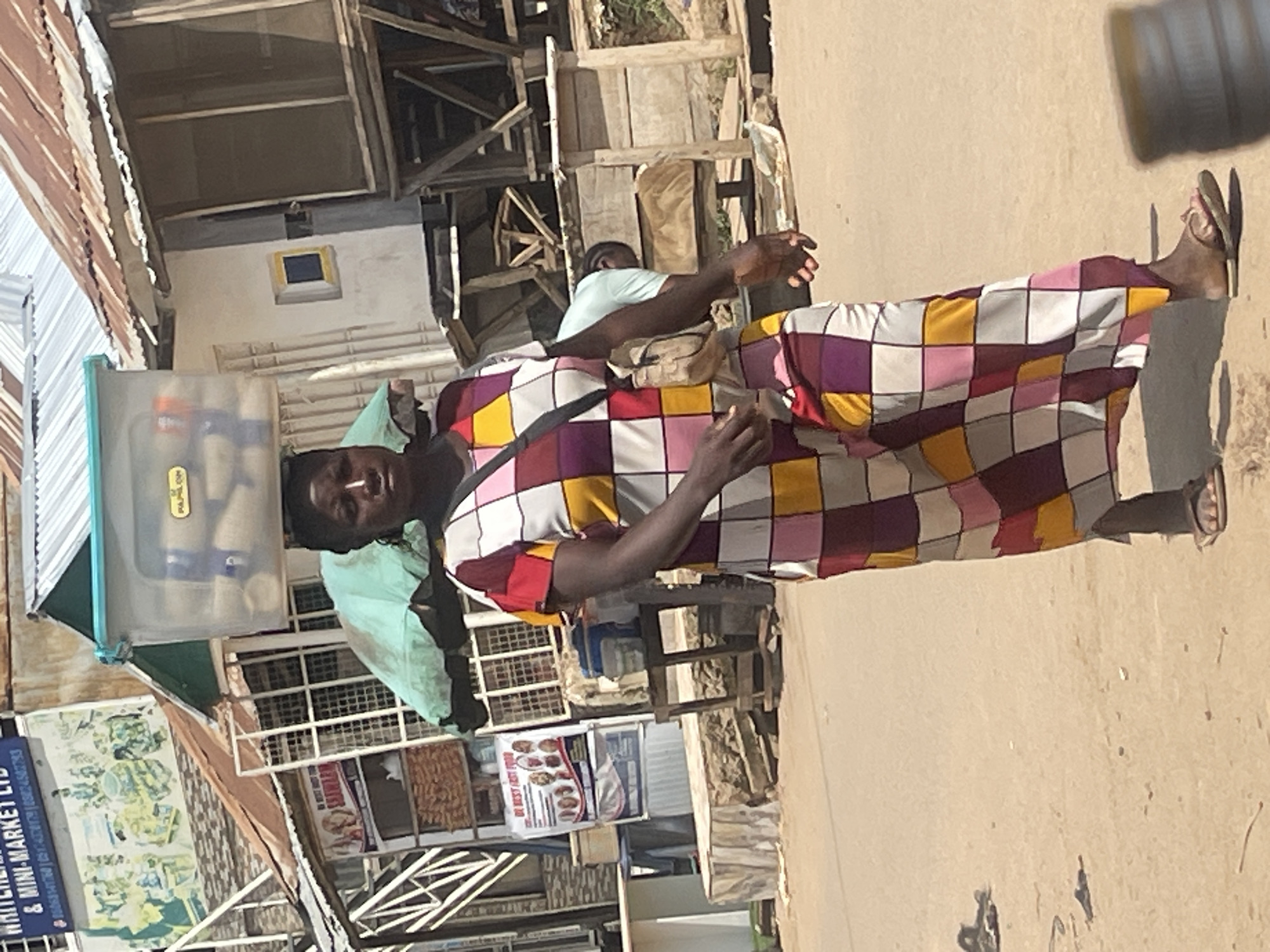 In the city of Abuja, Nigeria, a woman walks on a dirt road, carrying a clear plastic container filled with items on her head. She is wearing a long, colorful dress with a checkered pattern of maroon, yellow, pink, and white squares, and a small beige shoulder bag. She wears flip-flops. Behind her, there are various market stalls and buildings with corrugated tin roofs, some appearing weathered. A light green tarp is visible over some of the market goods. In the background, a man in a light blue t-shirt is visible behind some of the stalls. The ground is dusty and uneven, with some debris scattered on it. The sun appears to be shining, casting shadows.