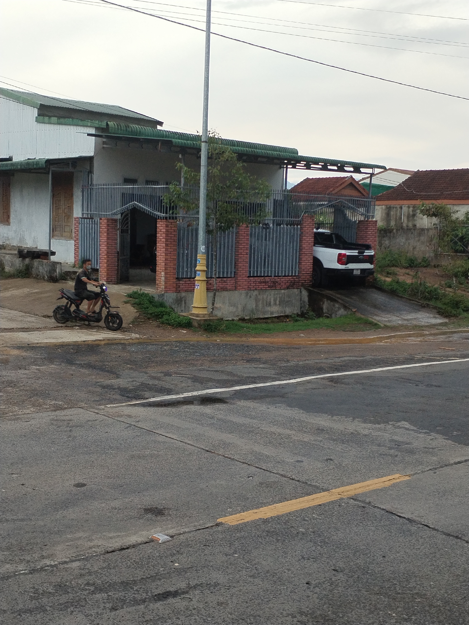A man wearing a mask is riding a small black electric scooter on a paved road in M'Drắk, Vietnam. In the background, a white pickup truck is parked in front of a house with a gated entrance. The house has a green roof and a combination of brick and gray metal fencing. A utility pole stands near the house. The road has some yellow markings and appears to be in moderate condition. The sky is overcast.
