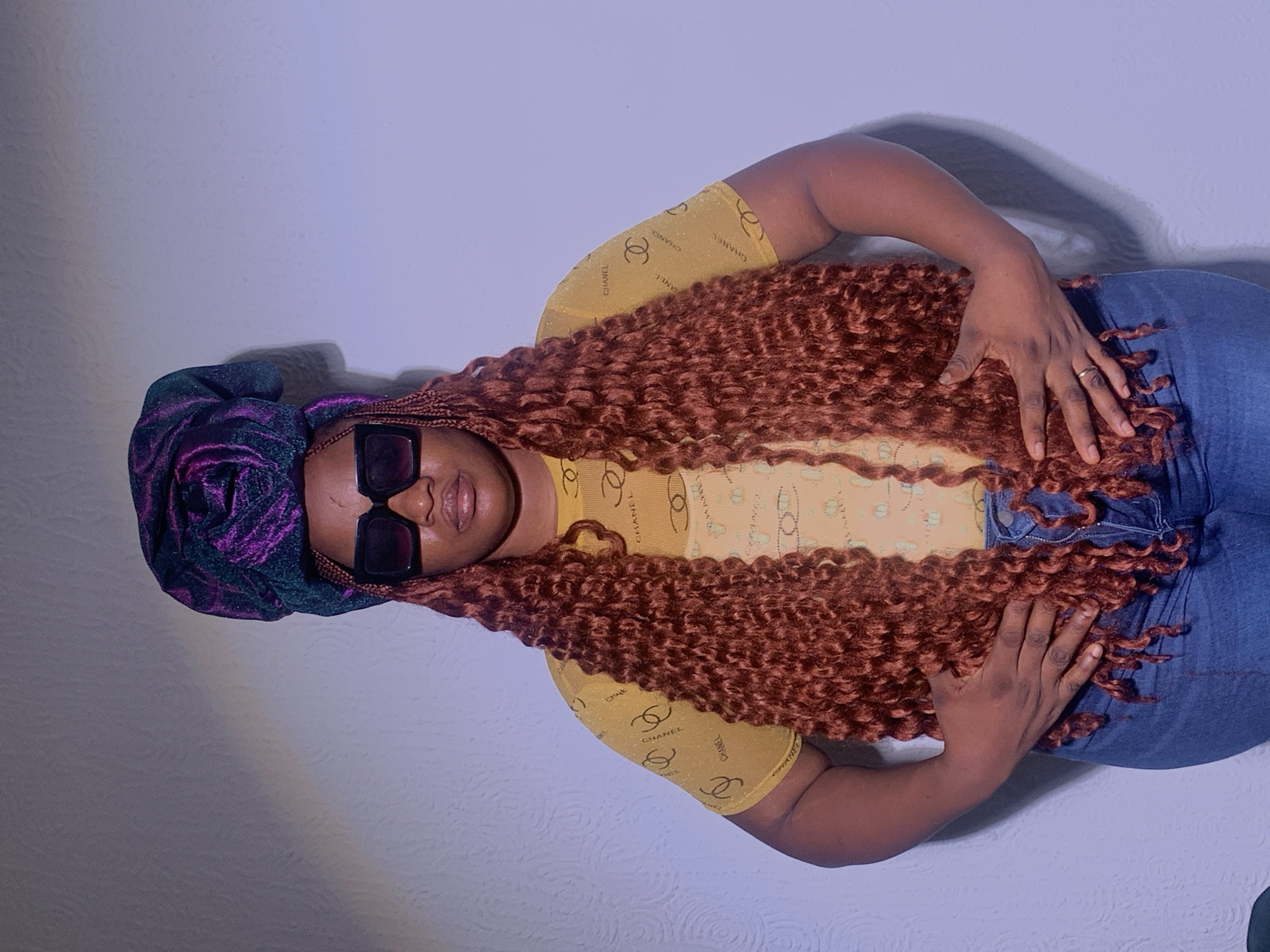 The image shows a woman lying on a light gray surface. She wears a dark head covering with purple and green patterns, large square sunglasses, a light yellow top patterned with the Chanel logo, and blue jeans. She has long, reddish-brown curly hair spread out across her torso. Her hands rest on her stomach and lap. The location is specified as Barakin Akawo, Nigeria, but there are no discernible environmental cues in the image to verify that location.