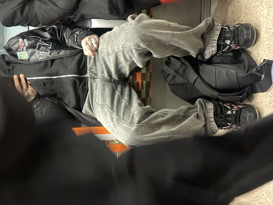 The image displays the interior of a London Underground train carriage, identifiable by the distinctive orange, brown, and green patterned moquette upholstery on the seats, placing the scene within the city of London, United Kingdom.
A person is seated centrally, occupying most of the frame from the torso downwards. They wear a black zipped hooded sweatshirt layered under a black, shiny bomber-style jacket featuring several patches and text, including "MP LUS" and "NAA." Their legs are clad in loose-fitting, light grey denim jeans with visible seam detailing and utility-style pockets. Black, chunky athletic shoes with white laces and red accents are on their feet. The individual's left hand, tattooed and adorned with rings, holds a dark-colored smartphone. A black fabric backpack is positioned on the speckled floor between their feet. To the right, a portion of another person seated next to the primary subject is visible, wearing dark clothing. The left side of the foreground is heavily obscured by a large, blurred dark mass, indicating the presence of another individual close to the camera.