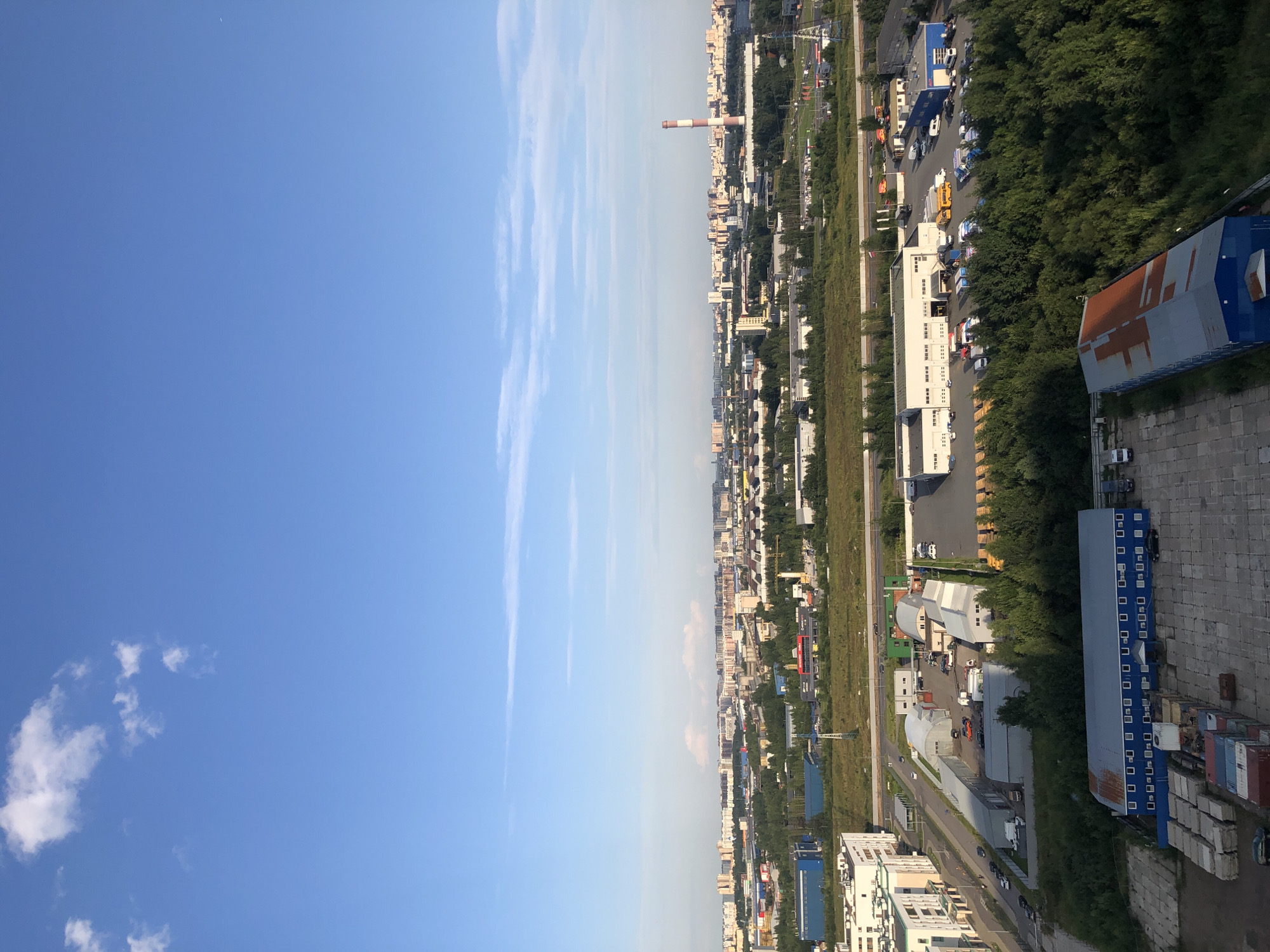 An aerial view captures a sprawling urban landscape under a bright blue sky with scattered white clouds. The foreground features a concrete lot with stacked concrete blocks and shipping containers, alongside a long blue building with a rusty metal roof and numerous windows. Further back, a dense green forest lines the edge of an industrial area. This area includes various warehouses and buildings, some with white facades and others with blue accents. A street runs between the industrial buildings, with several vehicles, including cars, trucks, and a yellow school bus, parked or in motion. Beyond the industrial zone, a wide expanse of residential buildings and urban infrastructure stretches towards the horizon. A tall, red and white striped smokestack stands out against the skyline, indicative of an industrial facility. The overall scene depicts a mixed urban and industrial environment on a clear day.
