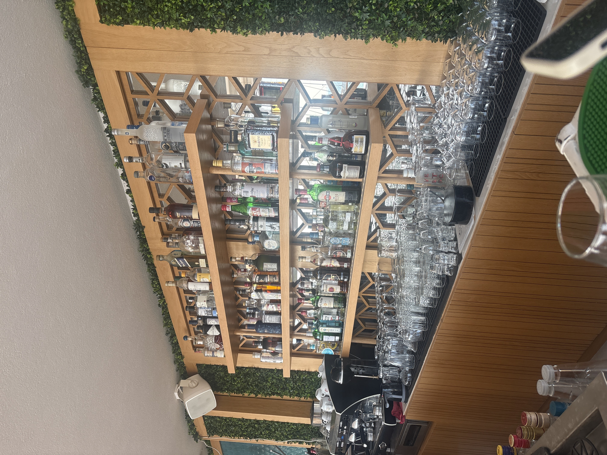 An indoor bar area features wooden shelving units stacked with numerous bottles of alcohol. The shelves are illuminated and the bottles display various labels and colors, suggesting a diverse selection of spirits. Below the shelves, a counter is filled with an assortment of clean glassware, including wine glasses and tumblers, arranged neatly on drying mats. To the left, a professional espresso machine is visible, along with several white cups. The background is decorated with artificial green foliage and a patterned screen element integrated into the shelving. The overall ambiance suggests a well-stocked and organized establishment, likely a restaurant or bar in Sithonia Municipal Unit, Greece, ready to serve patrons.