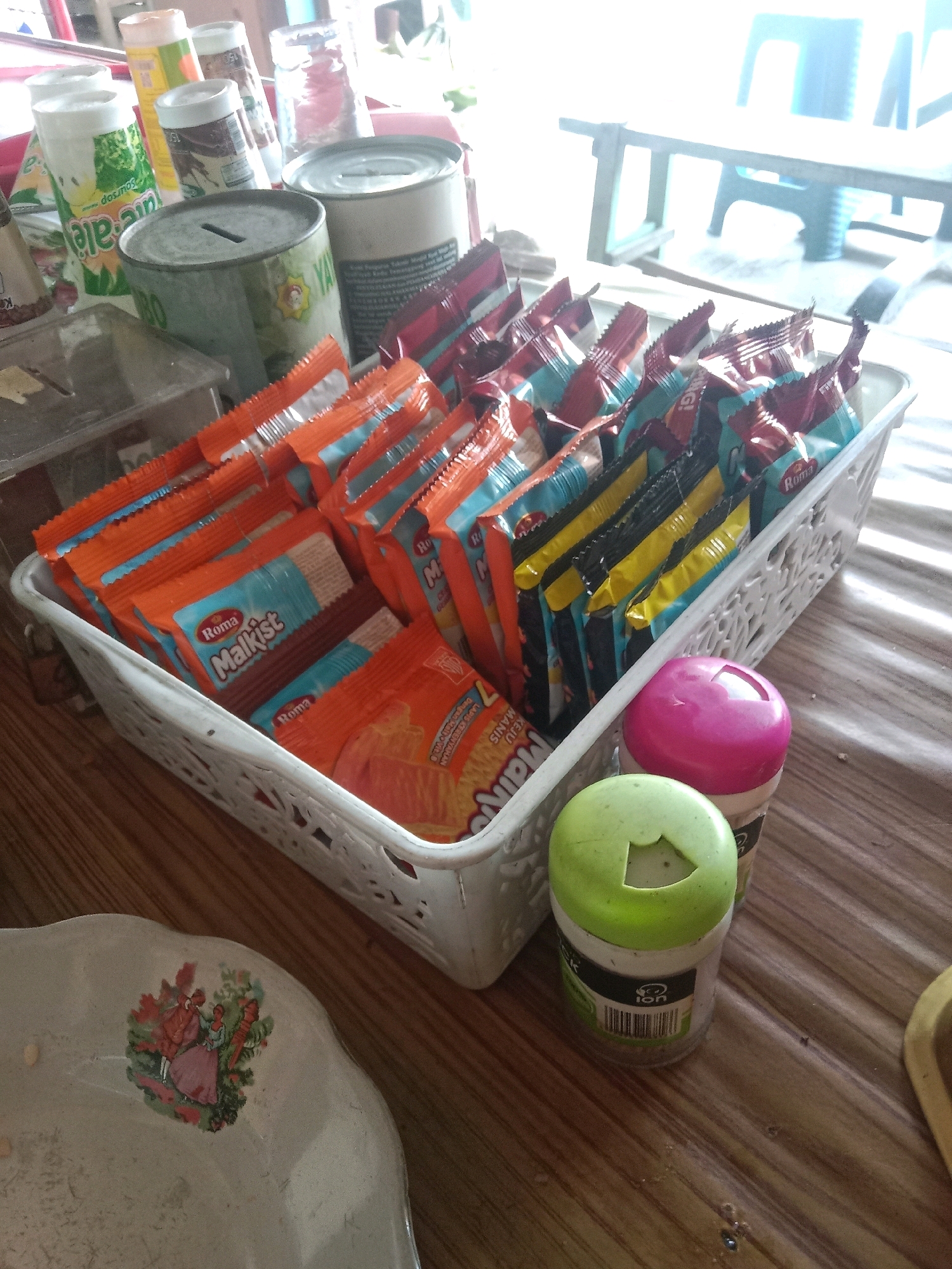 A basket of individually wrapped snacks, possibly cookies, is centrally placed on a wooden table in a room that appears to be a commercial establishment or home in Kertek, Indonesia. The basket contains multiple stacks of snacks, primarily in orange and blue packaging, with some brands like "Roma Malkist" visible. In front of the basket, two small containers with colorful lids, one pink and one green, are positioned on the table. To the left of the basket, a white ceramic plate with a decorative floral scene featuring two figures is partially visible. The background is blurred, showing glimpses of other items and furniture, suggesting an indoor setting. The lighting appears to be natural, possibly from a window or open space, creating a bright and slightly overexposed environment.