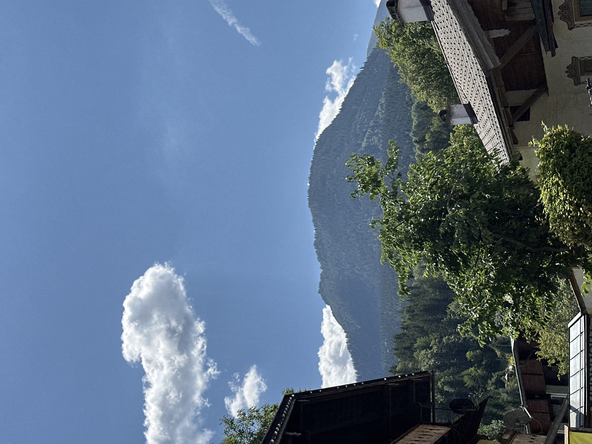 This is a photograph of the Watzmann mountain in Berchtesgaden, Germany, taken from a village street. The mountain is covered in lush green trees and has a large, imposing presence. The sky is a vibrant blue with a few fluffy white clouds scattered across it. In the foreground, there are several traditional Bavarian houses with their characteristic wooden balconies and tiled roofs. A large green tree with full foliage is in the middle ground, obscuring part of the view of the mountain. The overall impression is one of a picturesque mountain village on a sunny day.