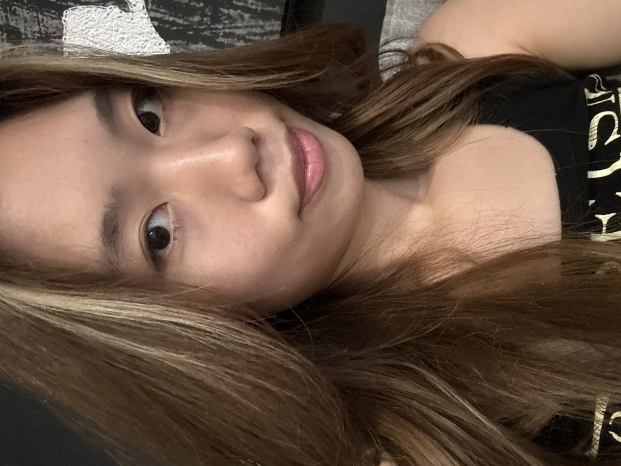 A close-up, slightly angled, and well-lit photograph depicts an East Asian female with fair skin. Her hair, a blend of brown and blonde highlights, is long and flows across the frame. She has dark eyes, a small nose, and pink lips. She is wearing a black top. The background is blurry and partially obscured by what appears to be a pillow or cushion with a black and white pattern, along with a gray textured surface. There is no information about this image being in Liangjiang, China.