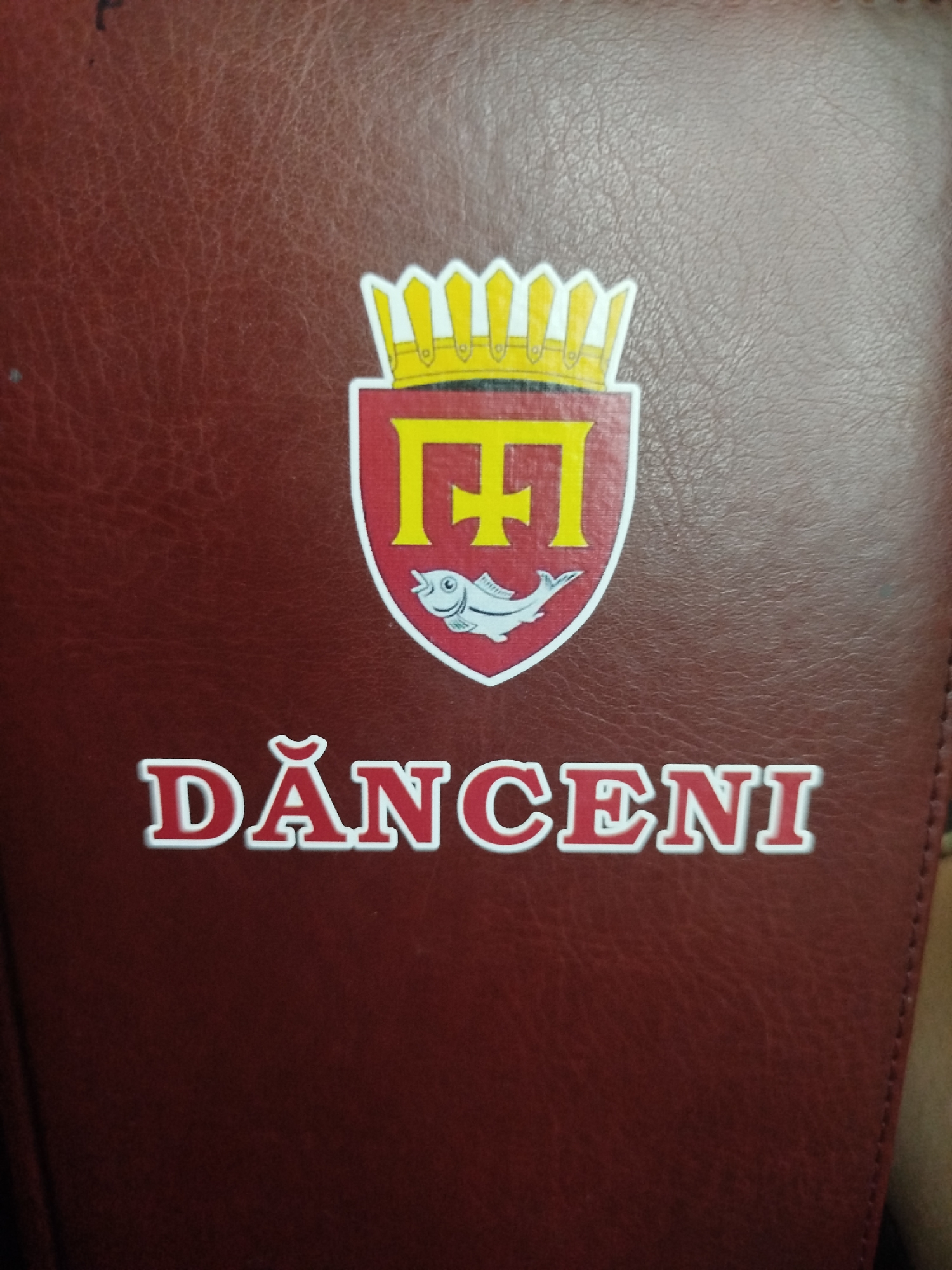 The image shows a close-up of a maroon, textured surface, possibly leather, bearing a coat of arms and text. The coat of arms is a shield with a crown at the top. Inside the shield, there is a stylized yellow letter "F" and a white fish. Below the coat of arms, the text "DĂNCENI" is prominently displayed in large, white, stylized letters with a red outline. The text and emblem appear to be a logo or insignia. The background is a plain, dark maroon color with visible texture. The overall impression is of a decorative element on a notebook, book cover, or similar item. The location in Nimoreni, Moldova, is not visually discernible from the image itself.