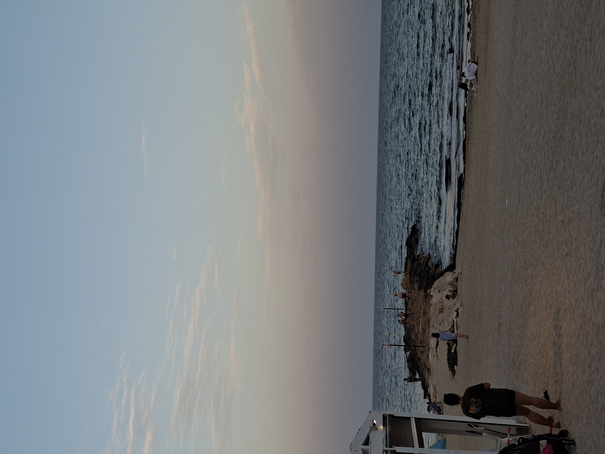 The image depicts a beach scene at dusk, likely in Torrevieja, Spain. The sky is a gradient of pale blue and soft pink, with wispy clouds suggesting the end of the day. The ocean is a deep blue with gentle waves lapping at the shore.
Several people are visible on the beach and near the water. In the foreground, a person is standing on the sand, facing away from the camera, with a stroller nearby. Further down the beach, other individuals are scattered, some near the water's edge and others on a rocky outcrop. One person in a white shirt is sitting by the water. There are also some surfboards or similar items visible near the water.
The sand is a light tan color, and the rocky area appears to be a natural part of the coastline. There are two posts with circular signs on them, though the text on the signs is not legible. A white structure, possibly a beach hut or changing room, is partially visible in the lower right corner. The overall atmosphere is peaceful and serene, indicative of a relaxing evening at the beach. The time of day is clearly evening, as evidenced by the sky and the soft lighting.