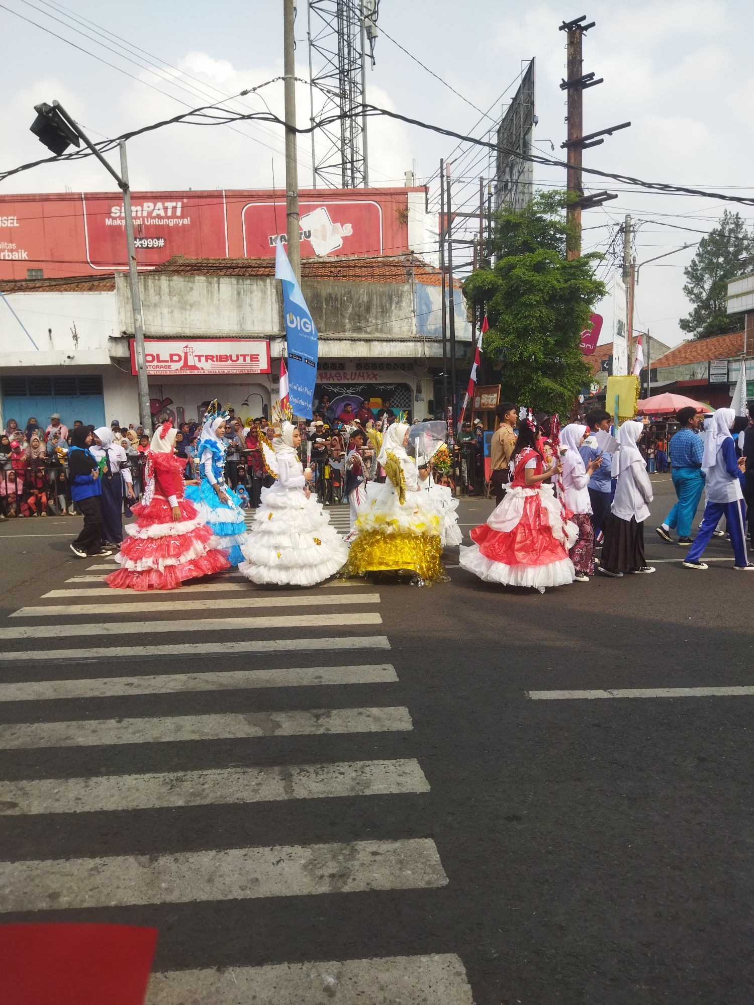 A street parade is in progress on a city street in Banjar, Indonesia. People in elaborate costumes, some resembling ruffled dresses in red, white, blue, and yellow, are walking across a pedestrian crosswalk. They appear to be participants in a cultural or festive event. A crowd of spectators lines the sidewalks on either side of the street, watching the procession. Buildings with signs, including "SinPati" and "OLD TRIBUTE," are visible in the background. Utility poles with numerous wires and a telecommunications tower are also present in the urban environment. The sky is cloudy.