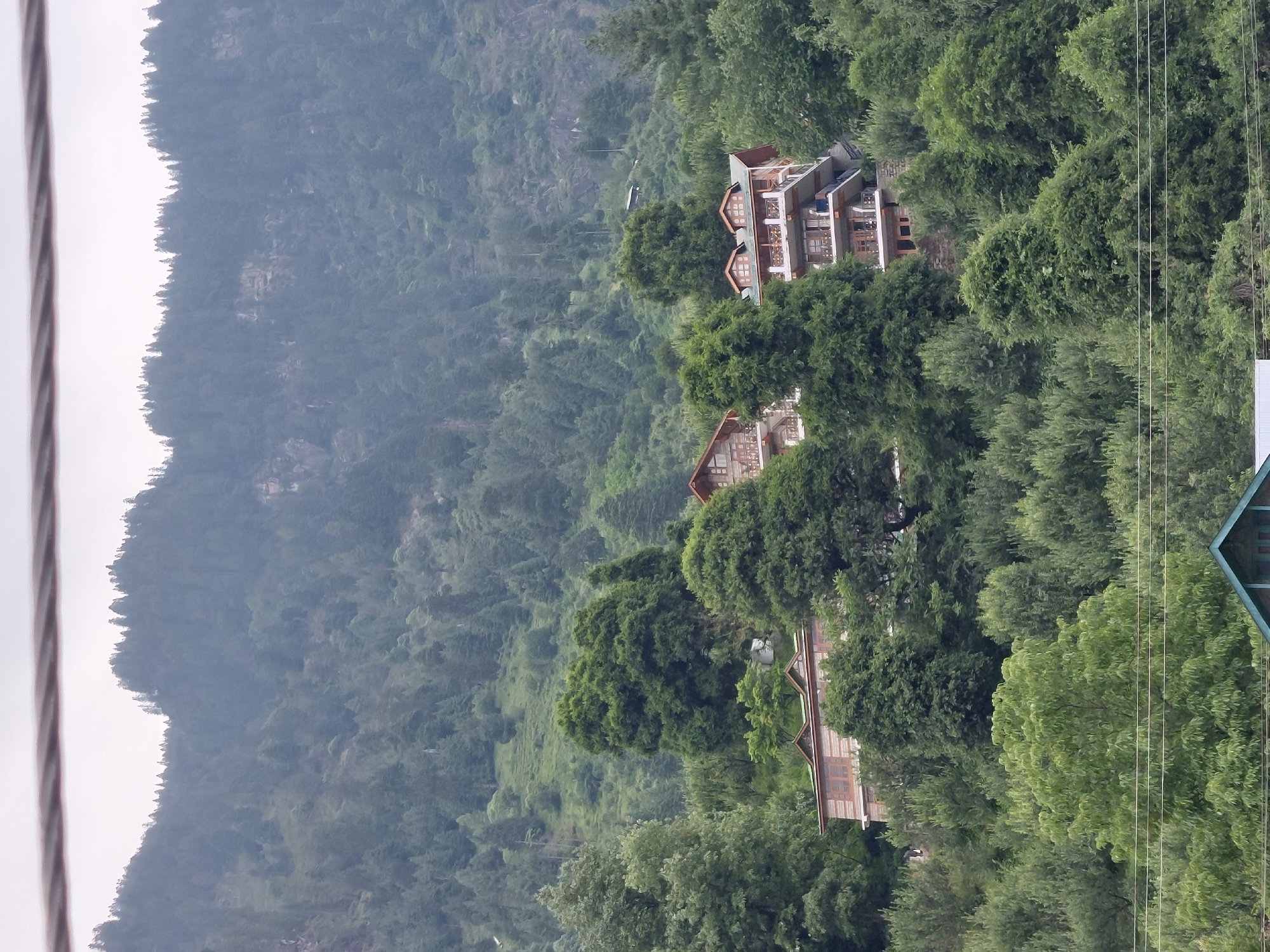 The image depicts a multi-story building nestled into a lush green mountainside, characteristic of Vashisht, India. The building features multiple balconies with wooden railings, traditional Indian architectural elements such as peaked roofs with dormer windows, and wooden paneling. It is surrounded by dense foliage and tall trees, with a mist-covered mountain range forming the backdrop. A visible power line stretches across the lower portion of the frame. The overall scene suggests a serene, natural setting, likely a hotel or residential property.