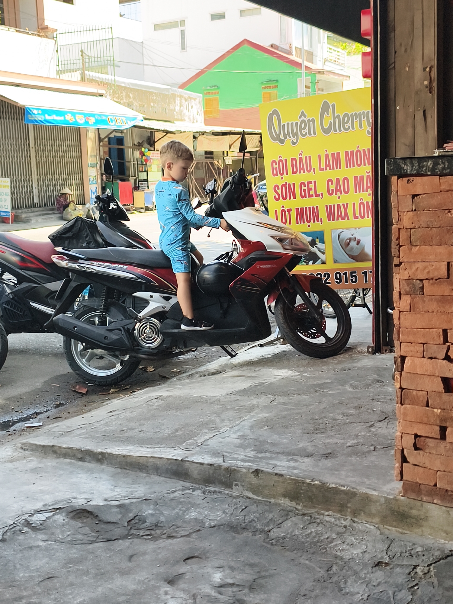 A young boy in a blue outfit is sitting on a red and white motorcycle parked on a concrete sidewalk. Another motorcycle is parked behind it. In the background, there's a yellow sign with Vietnamese writing, including the words "Quyên Cherry," advertising hair and beauty services. Behind the sign, there are buildings and structures typical of an urban setting in Bac Nha Trang Ward, Vietnam, where the scene is located. The day appears bright, with sunlight casting shadows on the street. A brick structure is visible on the right side of the frame.
