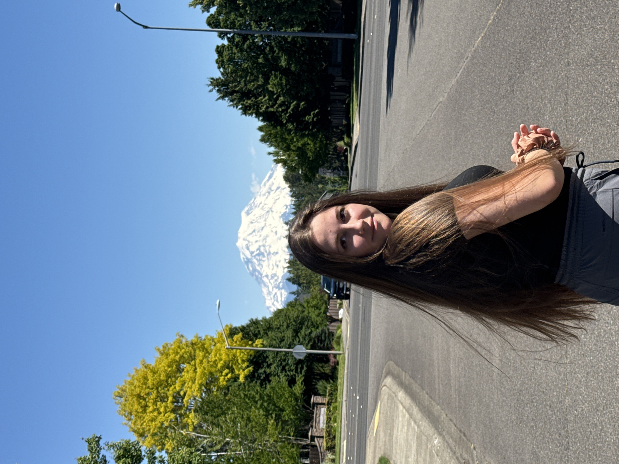 A young woman with long brown hair and a black t-shirt stands on a paved road in South Hill, USA, looking back towards the camera. Behind her, a snow-capped mountain, possibly Mount Rainier, rises majestically against a clear blue sky. Lush green trees line the street, with a bright yellow tree adding a splash of color to the scene. A street lamp and a stop sign are visible, along with a black car in the background. The woman is holding her hands clasped in front of her. The image captures a sunny day with ample daylight.