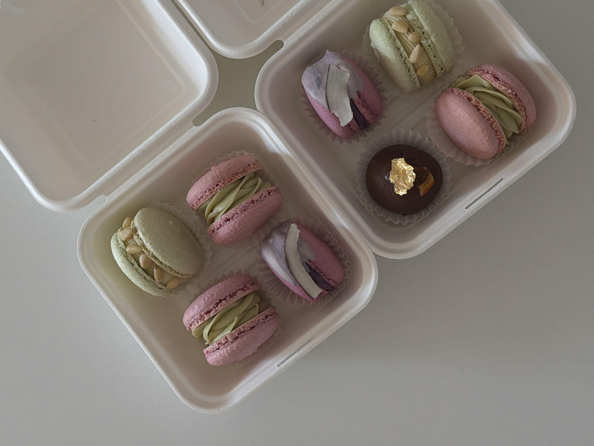 The image shows a top-down view of several macarons and a chocolate truffle arranged in two white take-out containers. The containers are placed on a plain white surface, which could be a table. The macarons are of various colors: pink, green, and purple. Some of the green macarons are topped with what appear to be nuts or seeds. The purple macarons have a decorative topping that resembles dried fruit or petals. The chocolate truffle is dark brown and has a piece of gold leaf on top. The overall lighting is soft and even, suggesting an indoor setting. There are no people or animals visible in the image, and no specific landmarks are depicted that would indicate a location in Tel-Aviv, Israel. The scene is static and appears to be a product shot or a still life arrangement.