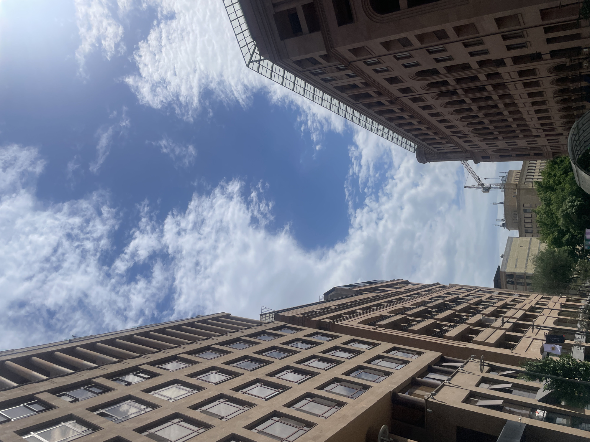 The image depicts a street scene in Yerevan, Armenia, with a view of two buildings and the sky. The buildings are tall, with multiple stories and numerous windows. One building has a glass-enclosed structure at its top. A construction crane is visible in the distance. The sky is blue with scattered white clouds. The angle of the image suggests it was taken from street level, looking upward. Some trees are visible at the right of the image, hinting at street-side vegetation.