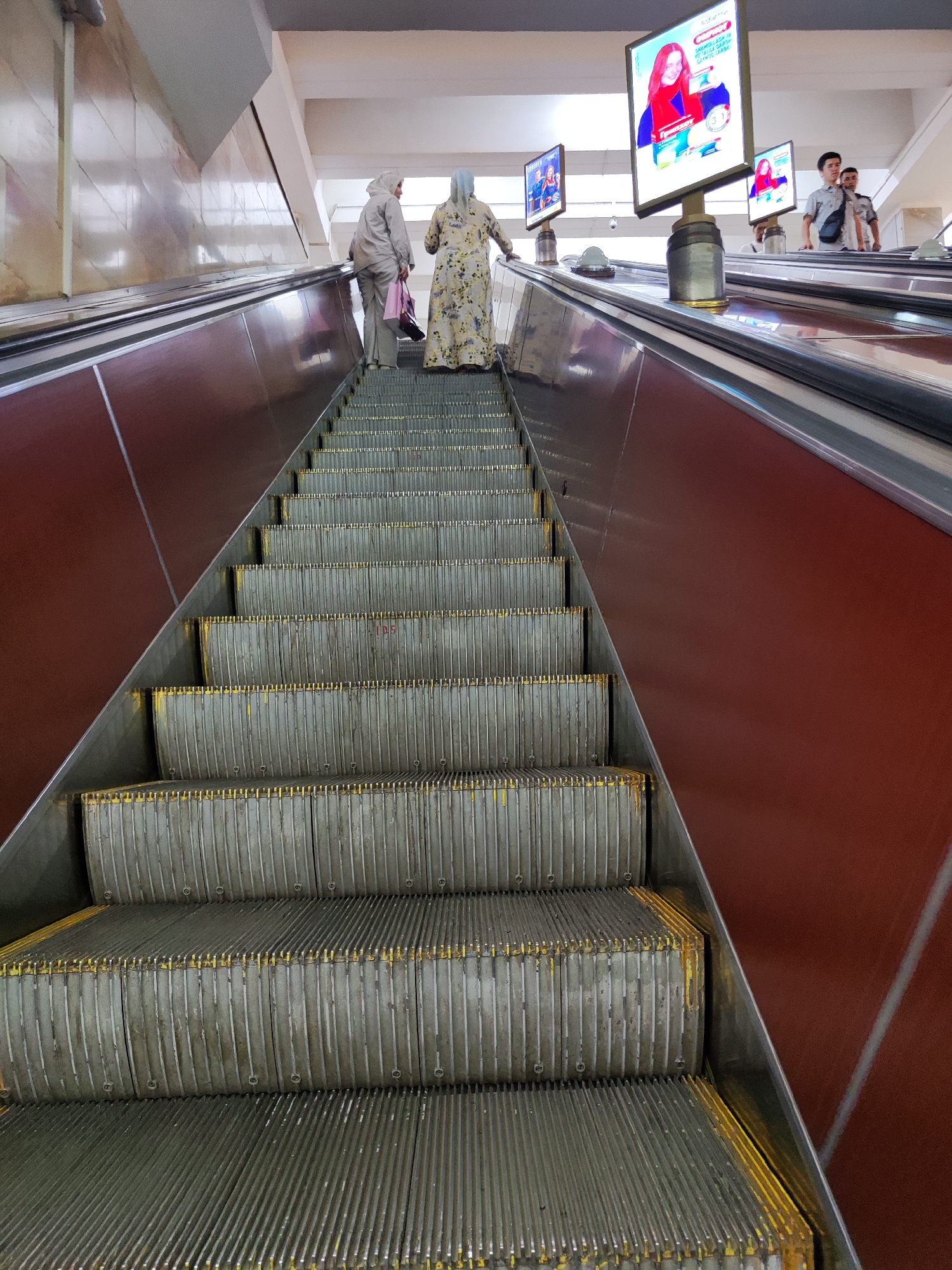 In Tashkent, Uzbekistan, two women ascend an escalator. The woman in front wears a floral-patterned dress and a light blue headscarf. The woman behind her wears a light grey tracksuit and a grey headscarf, carrying two shopping bags. They are on a wide escalator with metallic treads and a deep red side panel. Above them, three digital screens display advertisements. The furthest screen shows a woman holding a product, and the screen to its left shows two women. The nearest screen also displays an advertisement. Two men are visible further up the escalator, one in a light-colored shirt and the other in a dark shirt. The surrounding environment appears to be an indoor public space, possibly a metro station or a shopping mall, with a high ceiling and tiled walls. The lighting is bright and artificial.