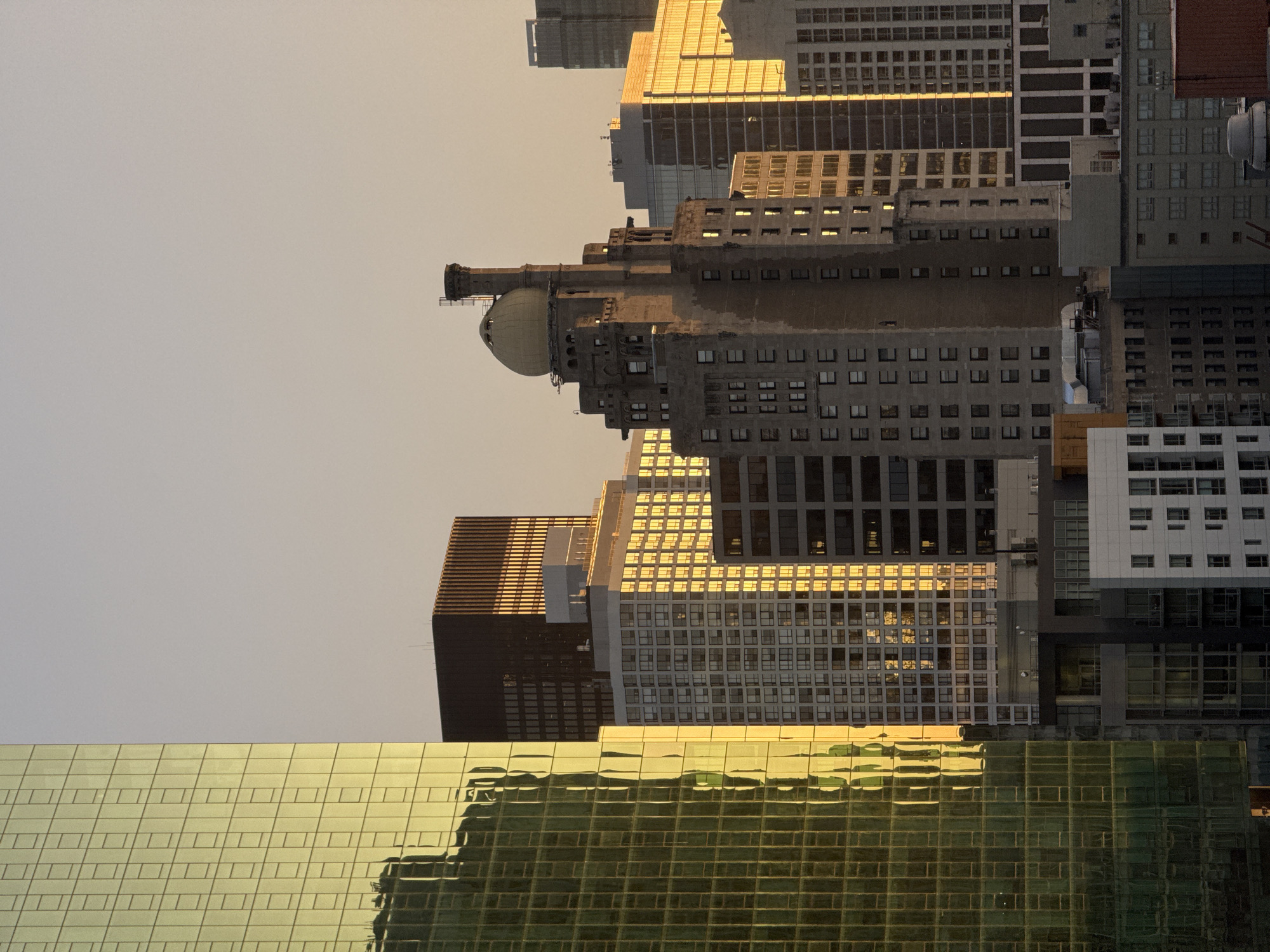 This is a close-up, medium shot, eye-level, outdoor, vertical, natural light photograph of a cityscape in Chicago, United States of America, at dusk or dawn. The image is dominated by various skyscrapers with a warm, golden light reflecting off their glass facades.
In the foreground and to the left is a towering building with a yellow, grid-patterned glass exterior. Its surface reflects the surrounding buildings and the sky in distorted, elongated shapes.
To the right of this building, and slightly behind it, is a dark brown, rectangular skyscraper with a ribbed facade. Sunlight illuminates the right side of this building.
Further to the right and slightly more prominent in the mid-ground is an older, tan-colored building with a distinctive dome-shaped structure on top, capped by a pointed spire. This building has numerous small, dark windows arranged in a repeating pattern.
Behind and to the right of this domed building are several other modern skyscrapers with glass exteriors, some appearing as a lighter shade of gold or yellow due to the sunlight. One of these buildings has a striped pattern on its facade.
The sky above is a pale, gradient of warm tones, suggesting either sunrise or sunset.
There are no people or vehicles visible in the frame. The scene is static and focused solely on the architecture. The environmental conditions appear clear and still, with the primary visual element being the interaction of light with the urban structures.