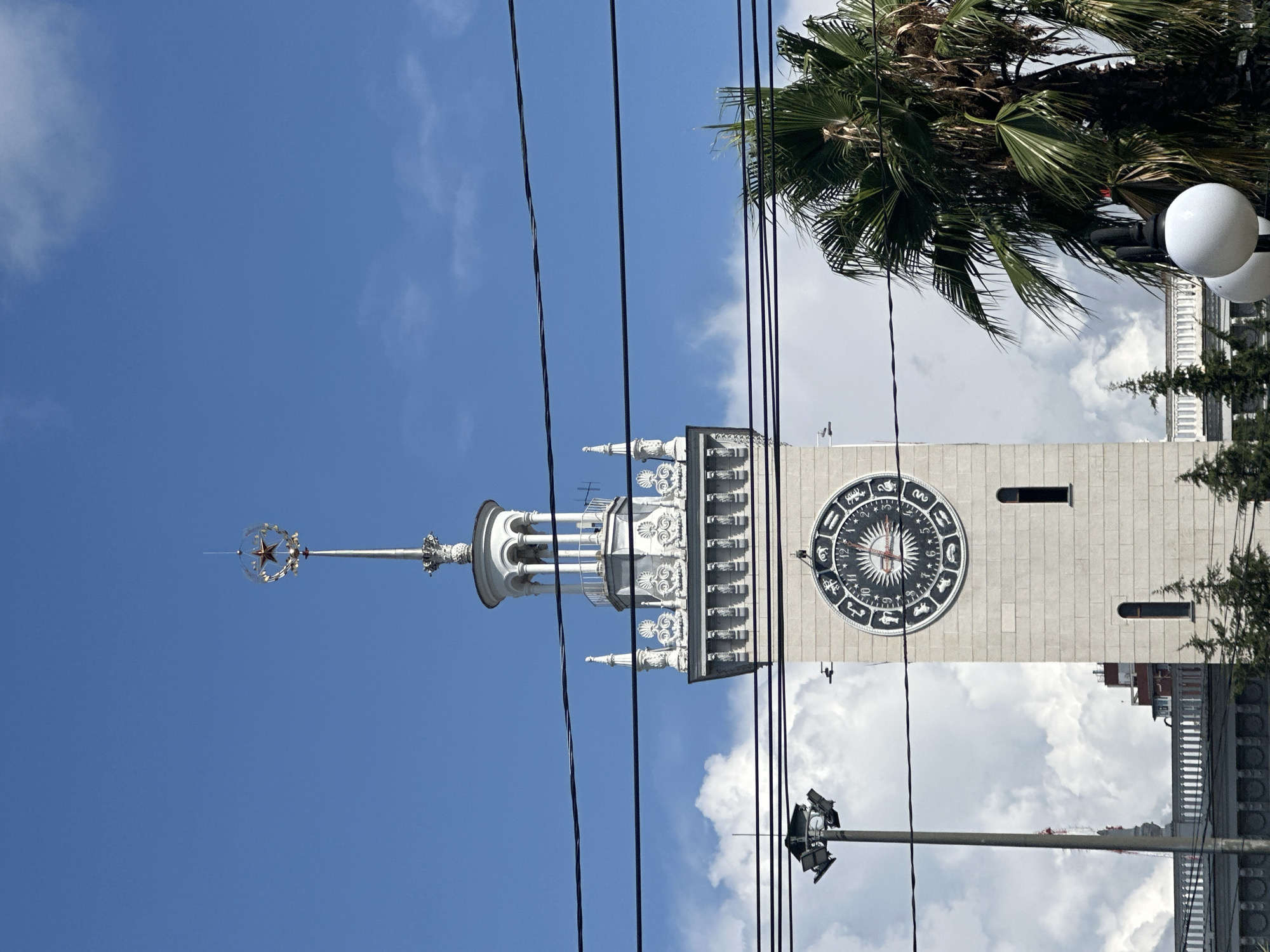 The image shows a close-up of the clock tower of Sochi's train station, a prominent landmark in the city of Sochi, Russia. The tower is made of light-colored stone and features intricate architectural details, including decorative carvings and a white ornate lantern-like structure at the top. A distinctive red star encased in a metallic sphere sits atop the spire. The clock face is large and round, with Roman numerals and zodiac symbols surrounding a central sunburst design. The hands of the clock indicate approximately 10:15.
In the foreground, several black electrical wires are strung across the frame, obscuring parts of the sky and the upper portion of the tower. To the left of the tower, a tall lamppost with multiple lights is visible. To the right, a large palm tree with lush green fronds partially obscures the tower. In the background, there are white, puffy clouds against a bright blue sky. Below the tower, the roofline of another building with a white balustrade and some evergreen trees are visible. The overall scene suggests a sunny day in a coastal city.