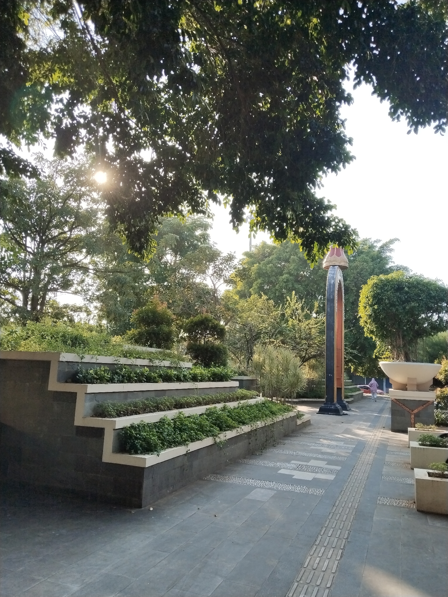 The image depicts a park with a paved walkway and a tiered planter box filled with greenery. A decorative archway with a finial stands to the right of the walkway, and a large planter with a white bowl-like structure is also visible. The scene is illuminated by sunlight filtering through the leaves of trees overhead. In the distance, a person in a pink headscarf and tunic is walking away from the camera. The environment appears to be a well-maintained urban green space.