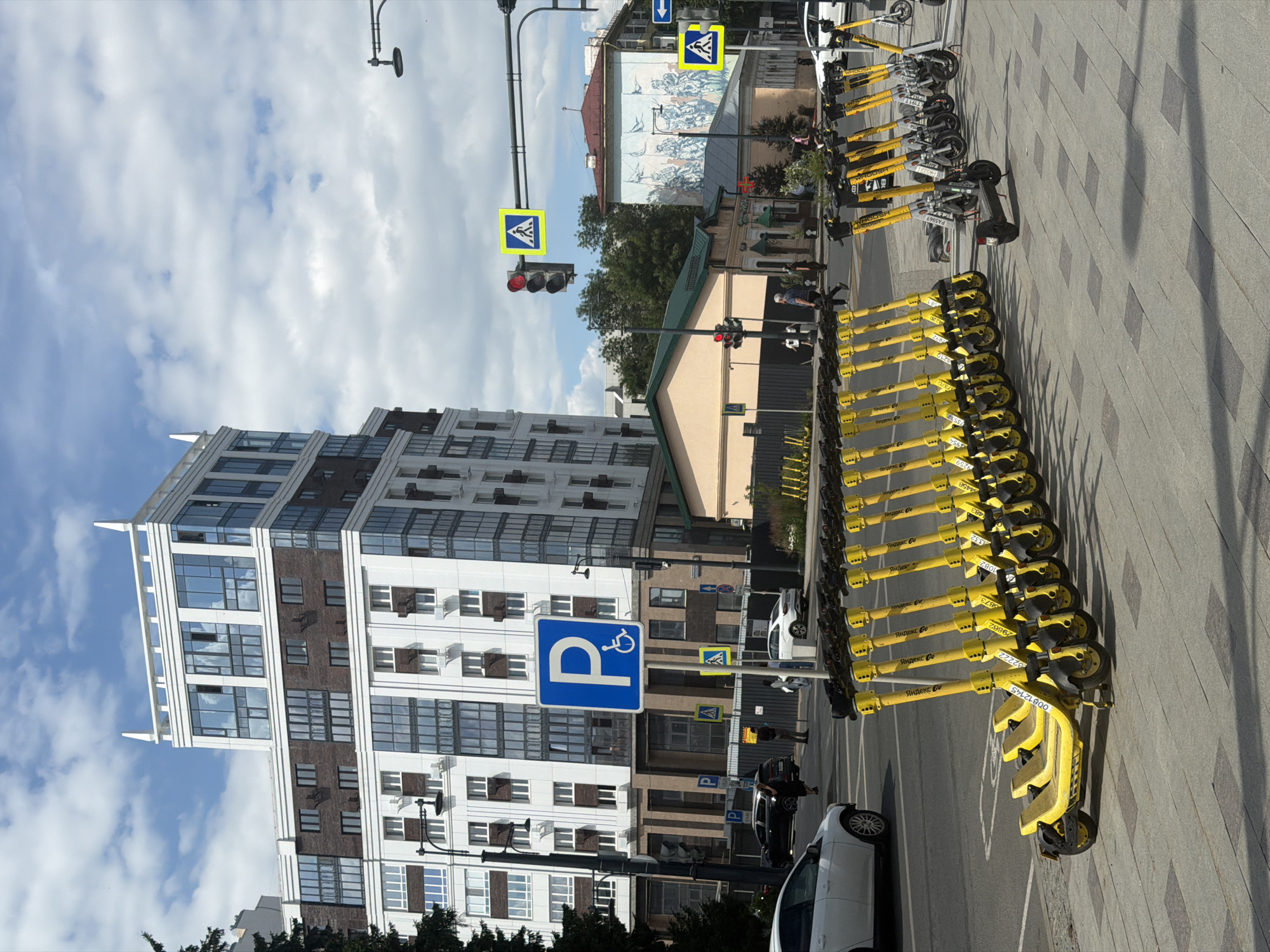 This vertical, eye-level, outdoor shot captures a city street scene on a cloudy day. The main subjects are a row of bright yellow electric scooters lined up neatly along a paved sidewalk. These scooters, branded with "Яндекс Go," appear to be available for rent. In the background, a modern, multi-story building with a mix of white and dark brick facades and numerous windows dominates the left side of the frame. Across the street, other buildings, including one with a mural depicting a battle scene, are visible. Traffic signals and pedestrian crossing signs are present, indicating an active intersection. Several cars, including a white one and a black one, are parked or driving on the road. A few people are walking along the sidewalk and crossing the street. The overall impression is one of a typical urban environment, likely in Ufa, Russia, given the visible text on the scooters. The cloudy sky suggests it might be late morning or afternoon.