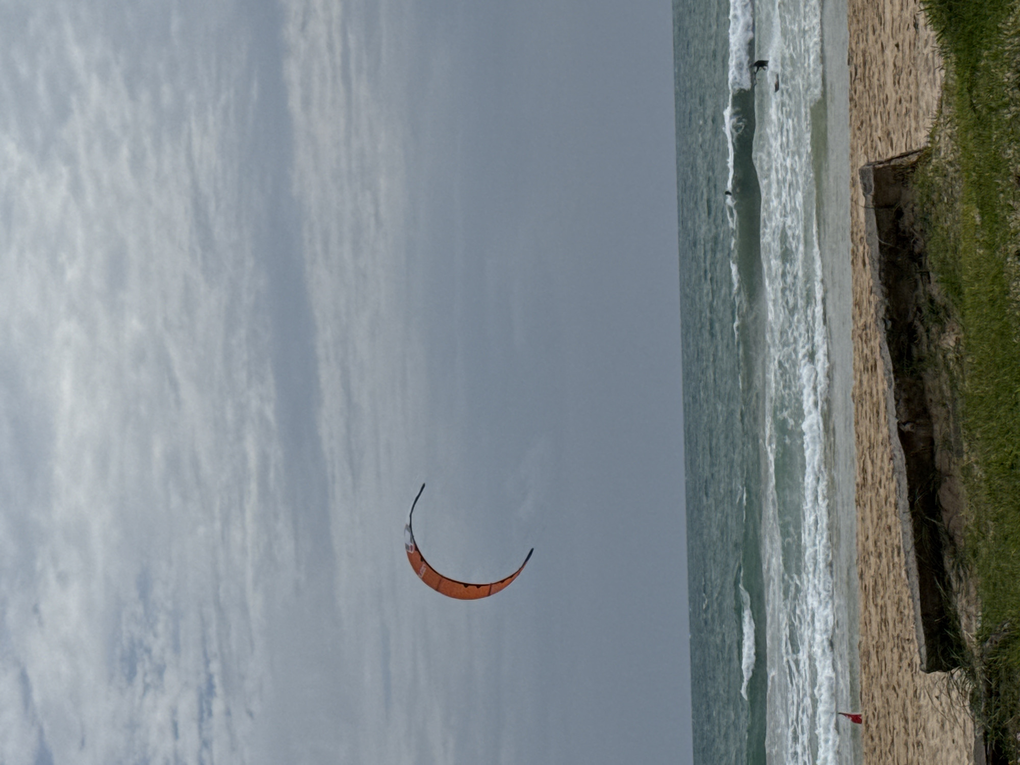 An overcast day at a beach in Rawai, Thailand. In the sky, a bright orange kite is visible against the gray clouds, curved like a crescent moon. The ocean is a choppy teal, with white waves breaking near the shore. On the beach, a red flag stands upright in the sand, and a large piece of driftwood lies on the sand near the foreground. Two surfers are visible in the water, one on a wave and another further out.