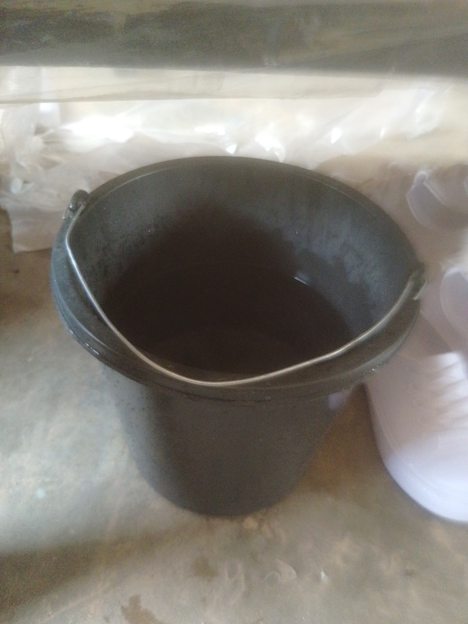 A black plastic bucket filled with water sits on a concrete floor. The bucket has a silver wire handle. Behind the bucket, a clear plastic sheet is draped over something out of focus. To the right of the bucket, a white plastic object with ridges is partially visible. The scene appears to be indoors or under a covered area, possibly outdoors given the concrete floor and the presence of what looks like a water spill or wetness on the floor. The lighting is somewhat dim, suggesting it's not direct sunlight. There is no visible text. The overall impression is one of a functional setting, perhaps for domestic use or a workshop. The image is taken from a slightly elevated angle, looking down into the bucket.
