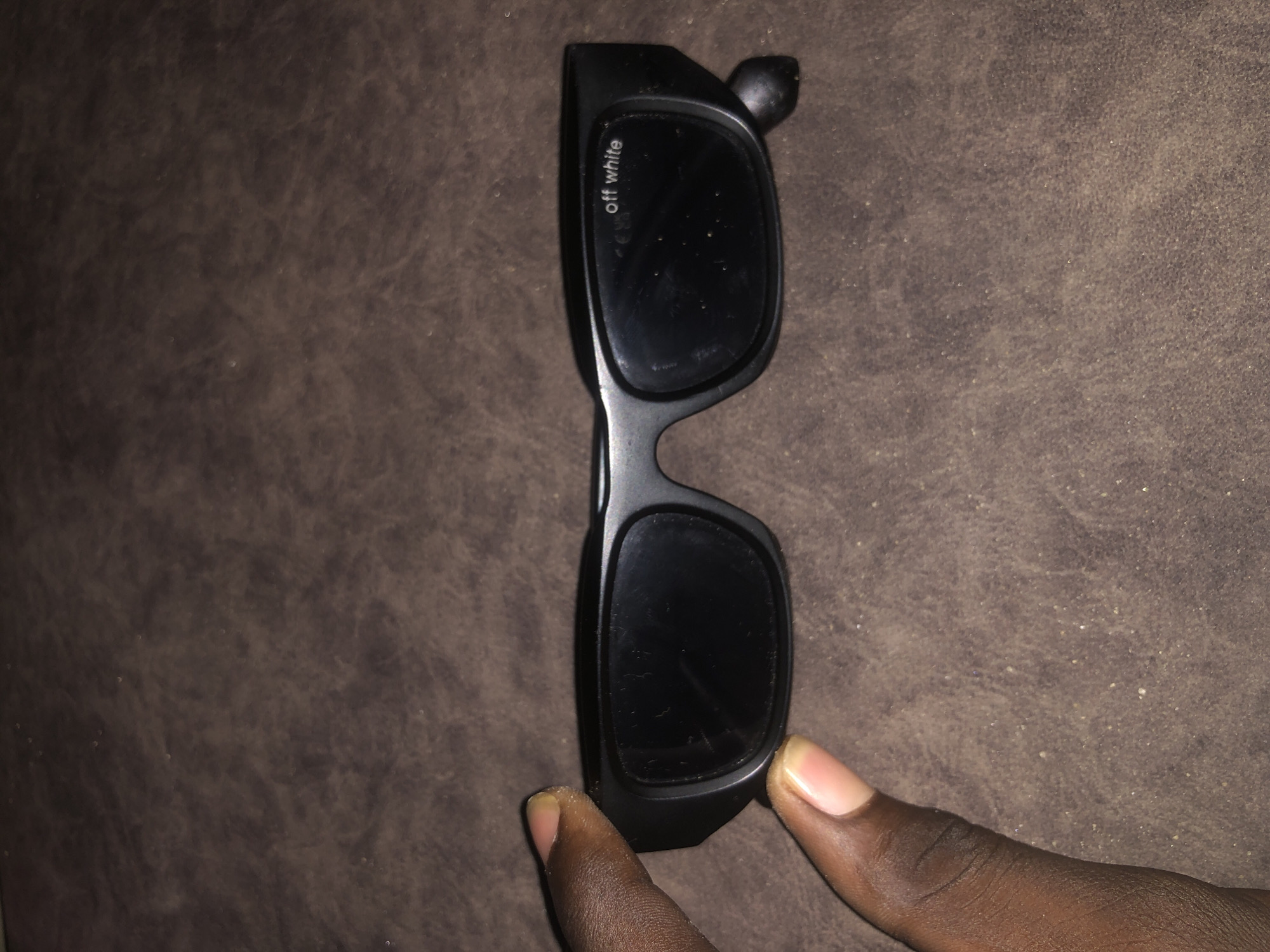 A pair of black sunglasses with the words "off white" printed on the right lens is held by a person's hand. The sunglasses are resting on a textured brown surface.