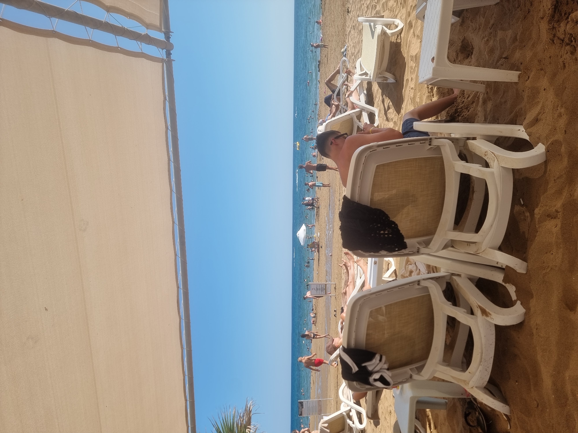 This is a photograph taken on a beach in Manavgat, Turkey. The scene depicts a bright, sunny day with a clear blue sky, suggesting pleasant weather for a beach outing. In the foreground, a beige sunshade partially obscures the view. The beach is populated with numerous people enjoying various activities.
Several white plastic lounge chairs are arranged on the sandy shore. Some are occupied by people relaxing, sunbathing, or resting. In the middle ground, a man is lying on a lounge chair, facing away from the camera. Further back, more beachgoers are visible, some walking along the shore, others wading in the calm, blue ocean. The water appears clear and inviting.
There are a few beach umbrellas scattered further down the beach, indicating a resort or public beach area. The overall atmosphere appears relaxed and leisurely, typical of a vacation setting. The time of day is likely midday or early afternoon, given the strong sunlight and the shadows cast by the chairs and people. No specific text is clearly legible within the image. The presence of the ocean, sand, and lounge chairs strongly points to a beach resort location.