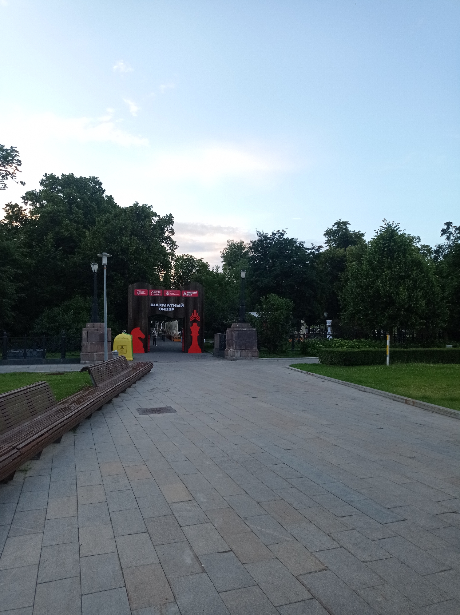 An outdoor shot shows a park entrance with the Russian text "ШАХМАТНЫЙ СКВЕР" (Chess Square) above a wooden archway adorned with chess piece figures. Two large red chess pieces, a knight and a queen, are positioned on either side of the entrance. A yellow trash bin is located to the left of the archway. The scene is illuminated by natural light, suggesting it's either dawn or dusk, with a clear sky overhead. A paved walkway leads towards the entrance, flanked by green grass and trees. Wooden benches are visible along the walkway in the foreground. The park appears to be a public space in Moscow, Russia, likely designed for leisure and recreational activities.