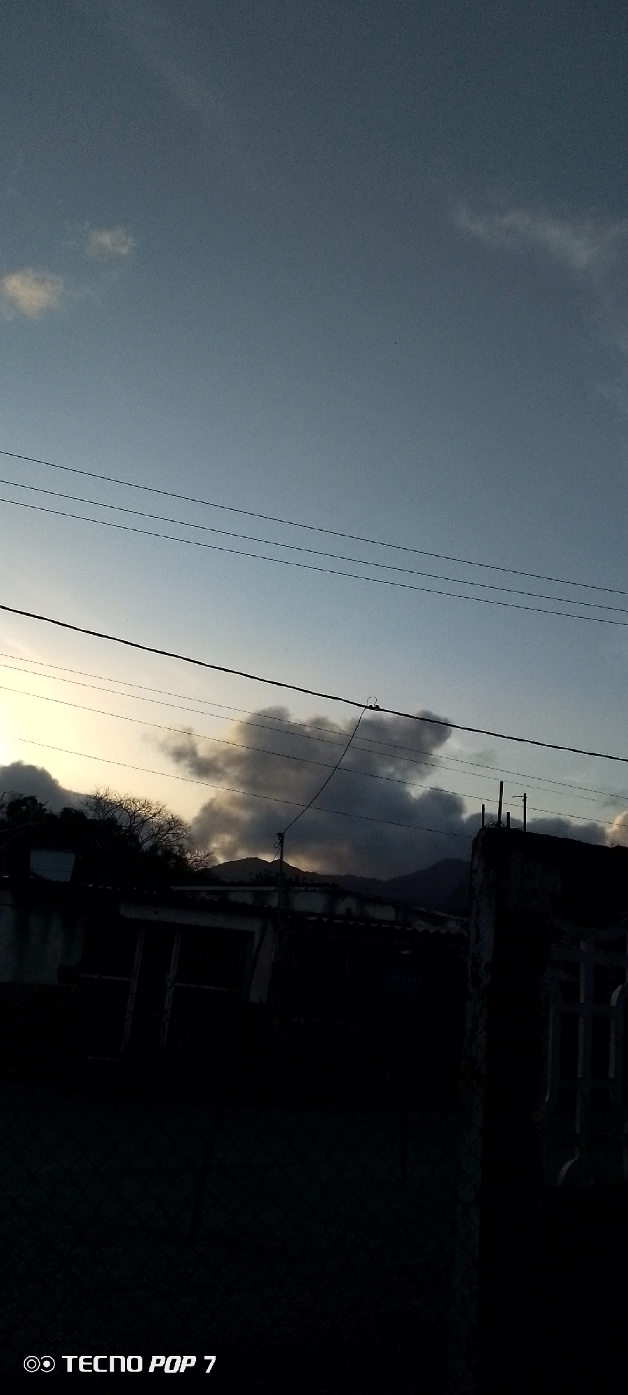 The image shows an outdoor scene in Altagracia, Venezuela, likely at dusk or dawn. The sky is a gradient of light blue to pale yellow/white near the horizon. Several dark clouds are present near the horizon, with some obscuring distant mountains. A series of electrical wires stretch across the upper portion of the frame. In the foreground, a silhouetted building is visible, along with parts of a fence on the right-hand side. The foreground is quite dark, making it difficult to discern details. The "TECNO POP 7" watermark is present in the lower left corner.