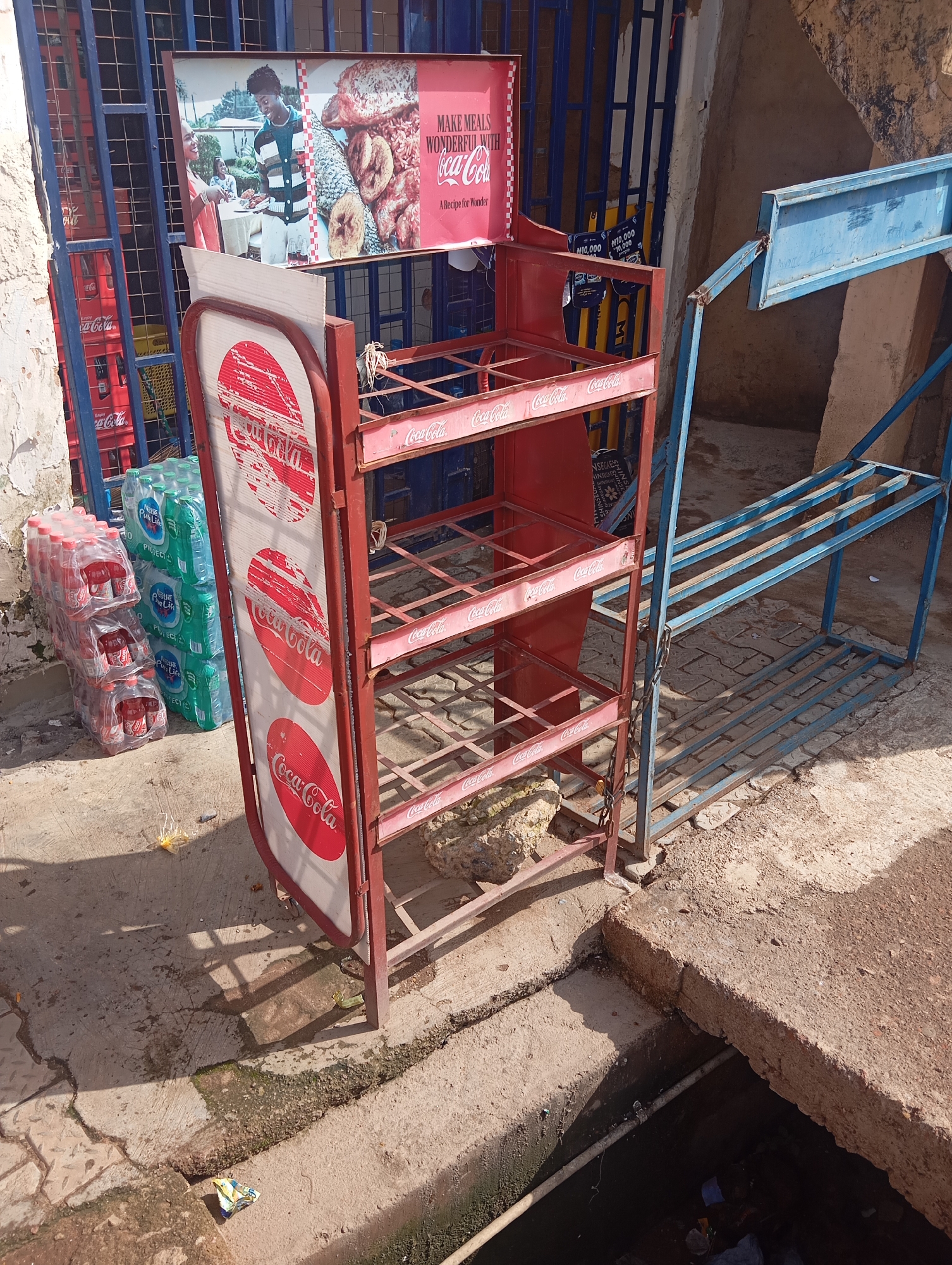 In Gura Top, Nigeria, a red metal Coca-Cola display rack stands on a concrete surface next to a blue metal shelving unit. Several cases of Coca-Cola bottles and green plastic bottles are stacked on the left side of the rack. A poster featuring a young couple, a young child, and various food items is attached to the top of the rack, with the text "MAKE MEALS WONDERFUL WITH Coca-Cola A Recipe for Wonder." The area appears to be an outdoor market or shop entrance, with a barred gate visible in the background. A narrow drain or gutter runs along the bottom of the scene.