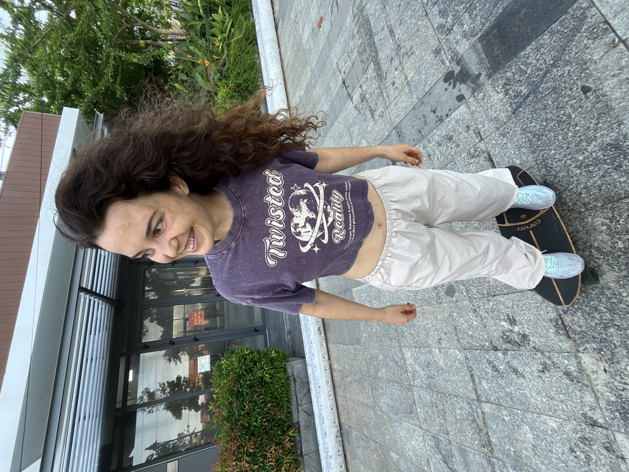 A young woman with long, curly brown hair stands on a skateboard on a paved walkway in front of a building with a modern facade. She is wearing a cropped purple t-shirt with a graphic print that says "Twisted Reality" and loose-fitting off-white pants. Her hair is blowing in the wind. The building has large glass windows and a covered entrance with horizontal louvers. Lush green bushes and trees are visible in the background. The ground is paved with large, grey granite tiles, and there is a white cylindrical object running along the edge of the walkway. The scene appears to be outdoors in a well-maintained urban environment. The image was taken in Phu Thuy Ward, Vietnam.