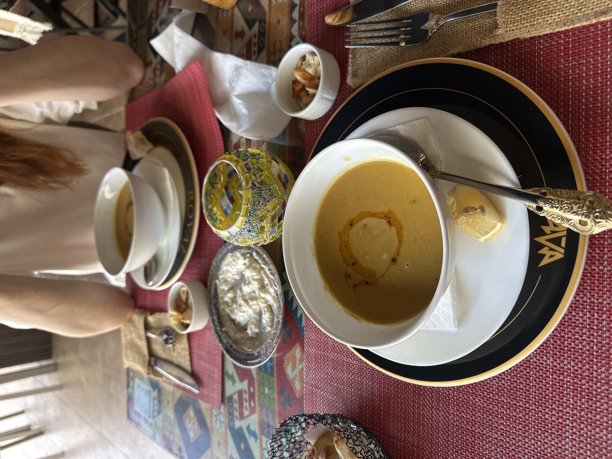 The image shows a table setting, possibly at a restaurant, with two place settings visible. A woman with long, light brown hair is partially visible on the left side of the frame. Each setting includes a bowl of yellow soup, a small dish of croutons, and a dish containing a creamy, white mixture (possibly tzatziki or a similar yogurt-based dip). There is also a decorative glass candle holder present. The right place setting has a fork and knife set resting on a burlap-textured napkin. The soup bowl on the right is placed on a plate with a black and gold rim. A lemon wedge is beside the soup bowl. The table is covered with a red textured tablecloth. The overall setting appears to be indoors. I am unable to determine the exact location or specifics regarding Baan Chaweng, Thailand based solely on this image.