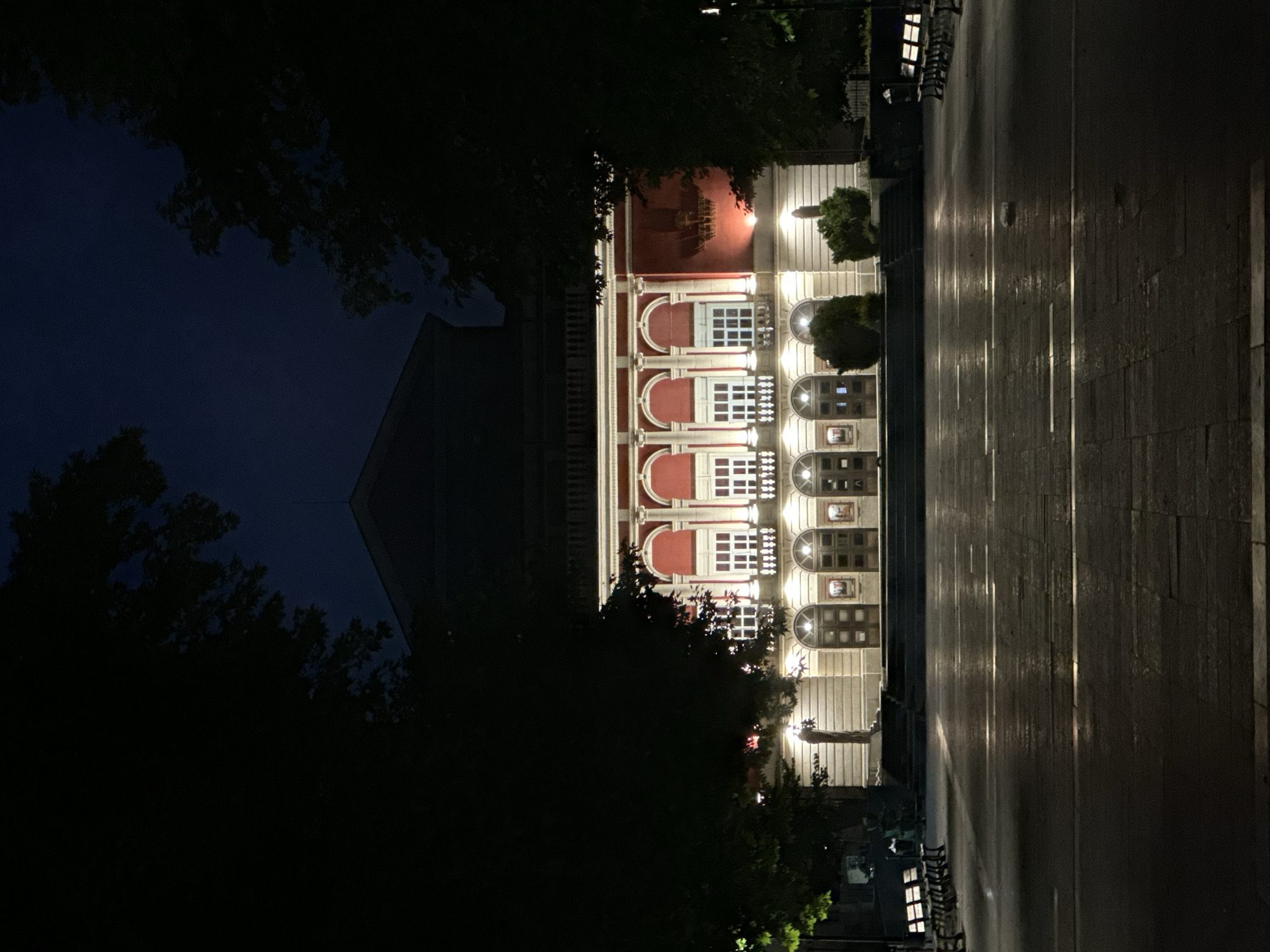 The image depicts the facade of the Ruse State Opera in Ruse, Bulgaria, at night. The building, a grand neoclassical structure, is illuminated by artificial lighting, highlighting its red and cream colored exterior. Arched windows and doorways are visible, along with ornate architectural details. The foreground features a paved plaza, wet and reflecting the lights, suggesting recent rain. Several benches are placed on either side of the plaza, and trees frame the scene, their dark foliage contrasting with the illuminated building. The sky above is dark, indicating nighttime. There are no people visible in the scene.