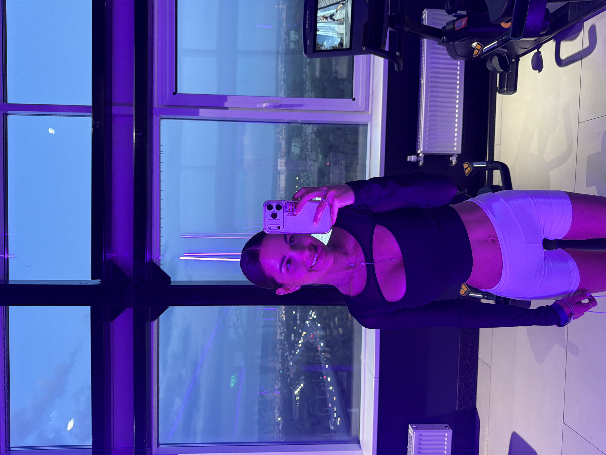 A woman is taking a selfie inside a building with large windows overlooking the city of Krasnoyarsk, Russia. The windows are sectioned by black metal beams and show a cityscape at dusk. The interior is illuminated with a purple-blue light. The woman is wearing a black long-sleeved top with a cut-out and white shorts. She is holding a smartphone in her hand, positioned to capture the image. In the background, exercise equipment is visible.