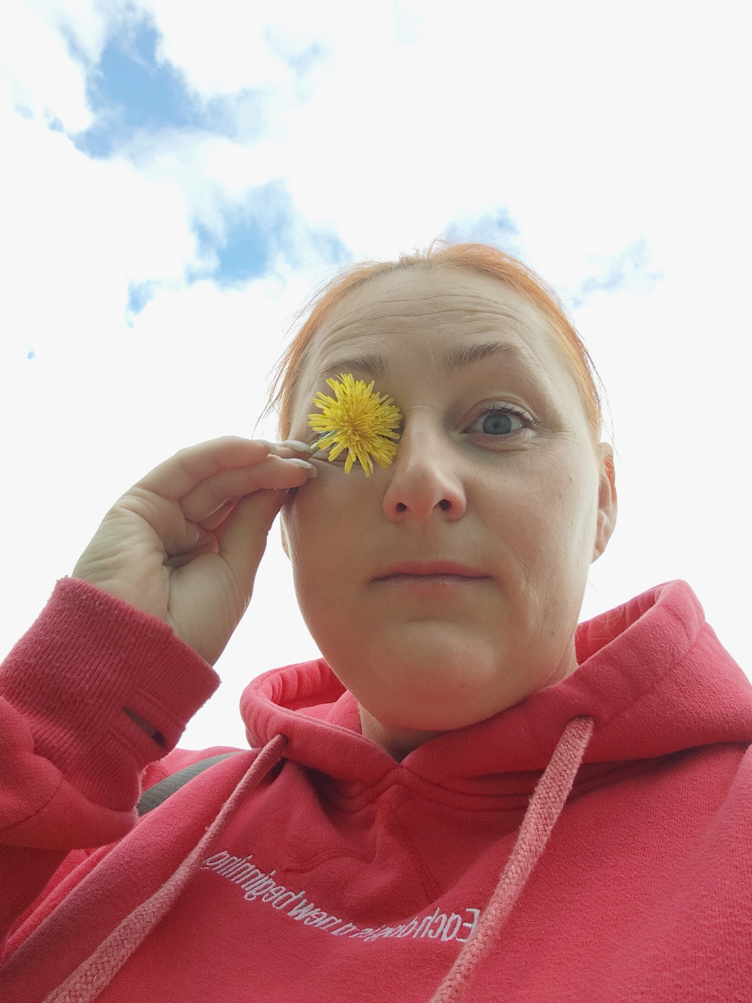 A woman with red hair, wearing a vibrant red hooded sweatshirt, is depicted in a low-angle shot looking up towards the sky. She is holding a bright yellow dandelion flower with her right hand, playfully covering her left eye. Her right eye is wide open and looking directly at the viewer. Her facial expression is neutral, possibly hinting at slight amusement. The background consists of a bright, clear sky with several scattered white clouds, indicating an outdoor setting during daytime.
Notable details include the texture of her red hoodie, which has visible drawstrings around the neck. Mirrored white text in Cyrillic script is visible on the front of her sweatshirt, appearing to say "приобретенный" or "приобретение" (meaning "acquired" or "acquisition"). No specific geographical landmarks are visible within the image to confirm the location, but the provided context is Markovskii, Russia. The sunlight suggests good weather conditions.