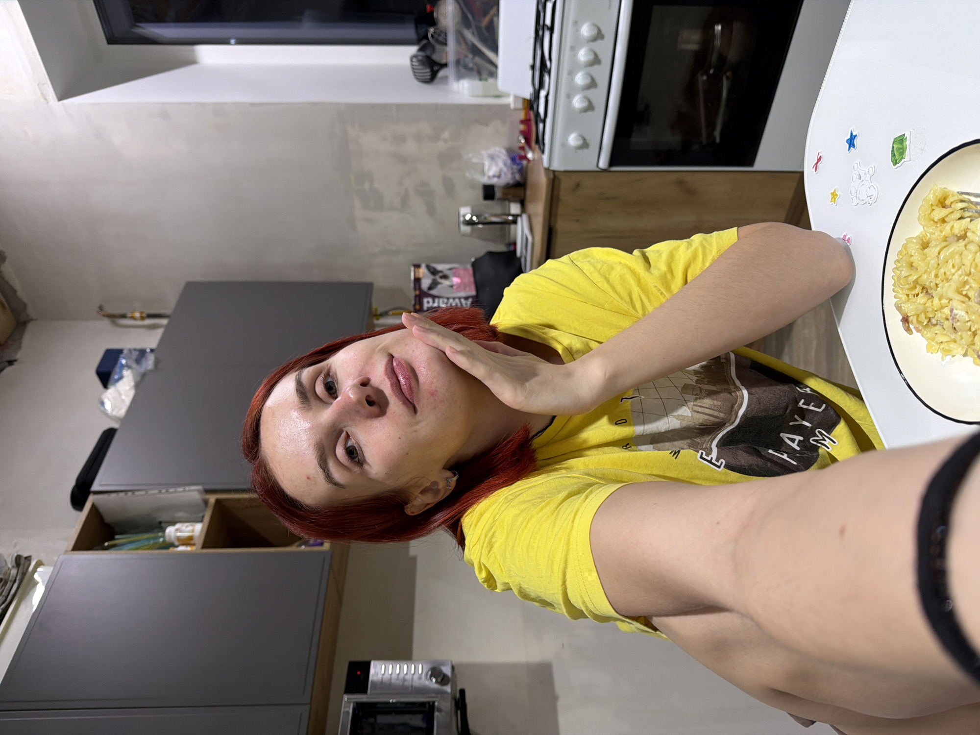 A woman with red hair, wearing a yellow t-shirt, is posing for a selfie in a kitchen. She is holding her hand to her face. In front of her is a plate of pasta. Behind her are kitchen cabinets, an oven, and a countertop with various items on it. Based on the available details, the image seems to be a casual personal photograph taken indoors. The location within Chaikovsky, Russia, is not evident from the image.