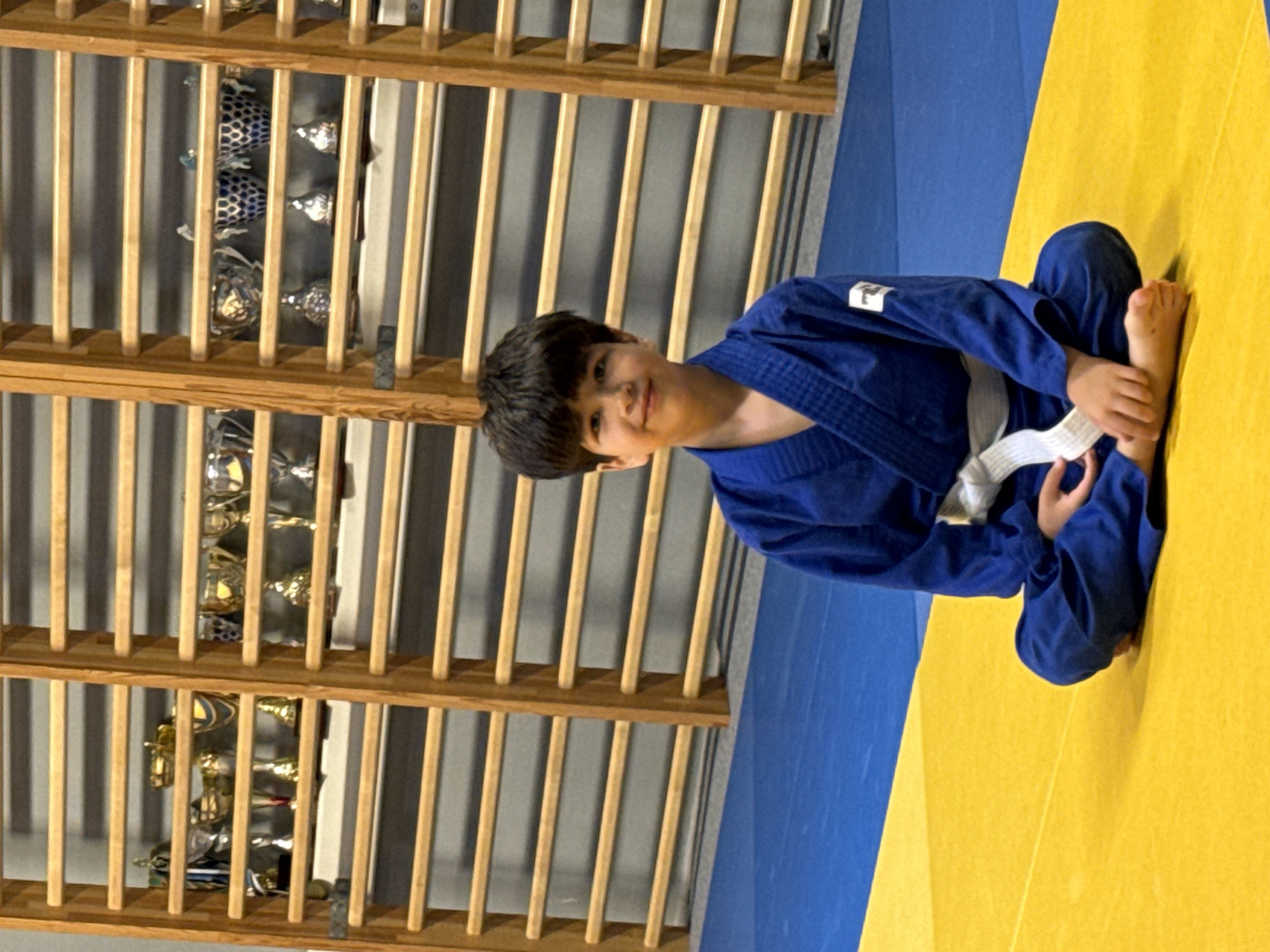 A young boy in a blue judo uniform with a white belt is sitting cross-legged on a yellow mat. The mat is bordered by a blue mat. Behind the boy, there are wooden shelves filled with trophies and awards. The shelves are part of a wall-mounted structure, possibly a gymnasium or training facility. The boy is looking towards the camera with a slight smile.