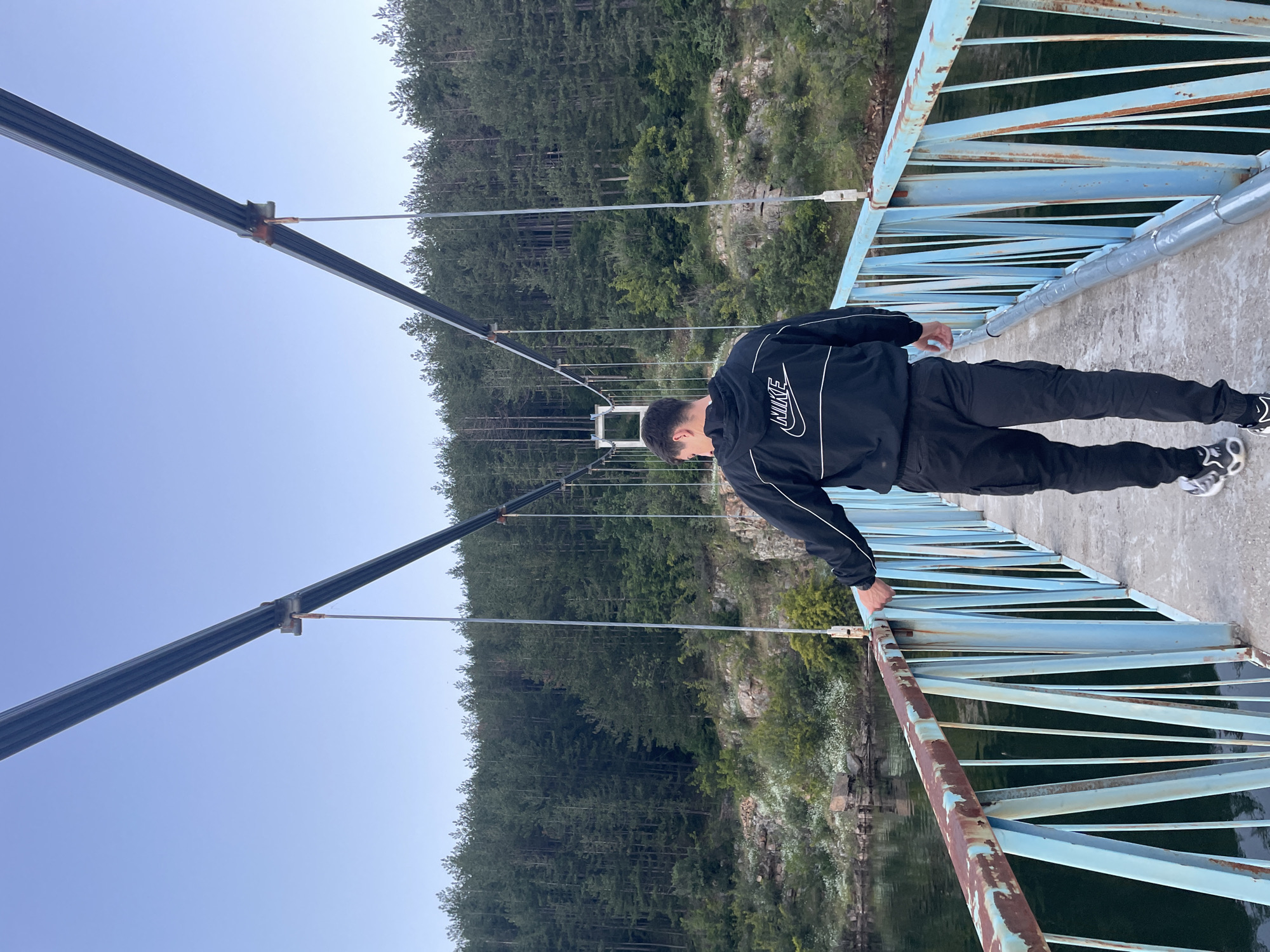 In the city of Suhindol, Bulgaria, a young man is walking across a suspension bridge. The man is wearing a black Nike tracksuit and black and white sneakers. He is walking away from the camera, towards a dense forest that covers the hills on the other side of a body of water. The bridge has a light blue metal railing with some rust spots, and the deck is made of concrete. The sky is clear and blue.