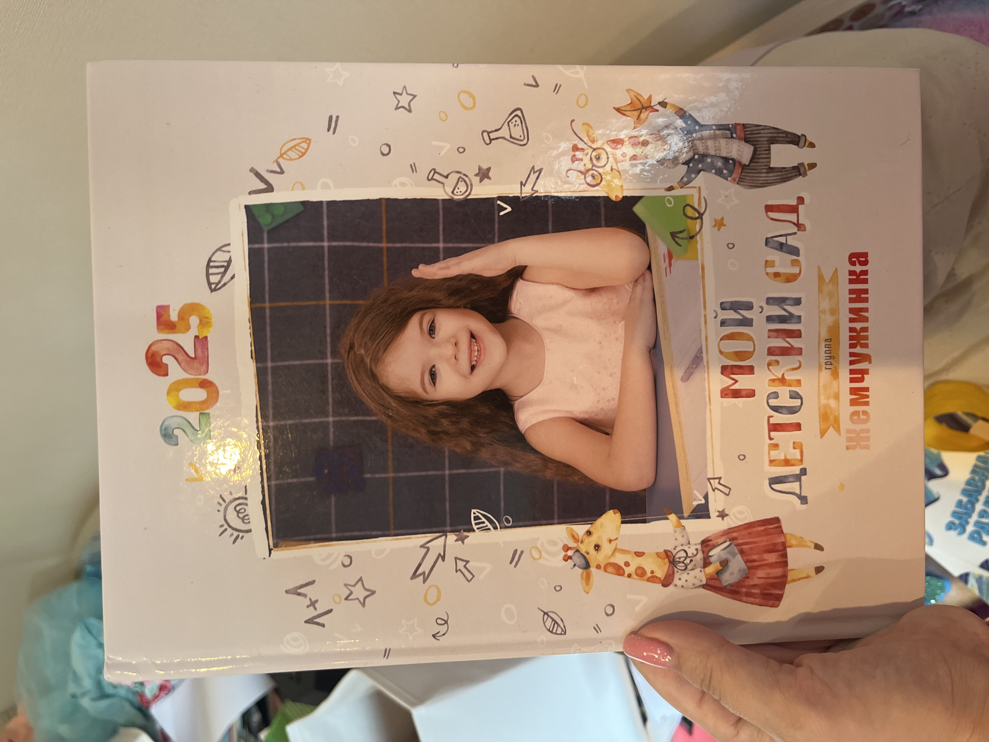 The image shows a close-up of a children's book cover. The book is white with colorful illustrations and text. A photograph of a young girl with long, wavy brown hair is in the center, framed by drawings of a chalkboard. The girl is smiling and has her right hand raised. The book's title, "МОЙ ДЕТСКИЙ САД" (My Kindergarten), is printed in large, colorful letters below the photograph. The text "2025" is at the top of the book, and below the title, it reads "группа Жемчужинка" (Group Pearl). There are also illustrations of giraffes, leaves, and other playful elements scattered around the cover. The book is held by a person's hand with pink nail polish. The background is blurred, showing a light-colored wall and possibly some household items. There is no indication of the city of Sochi, Russia, in the image.