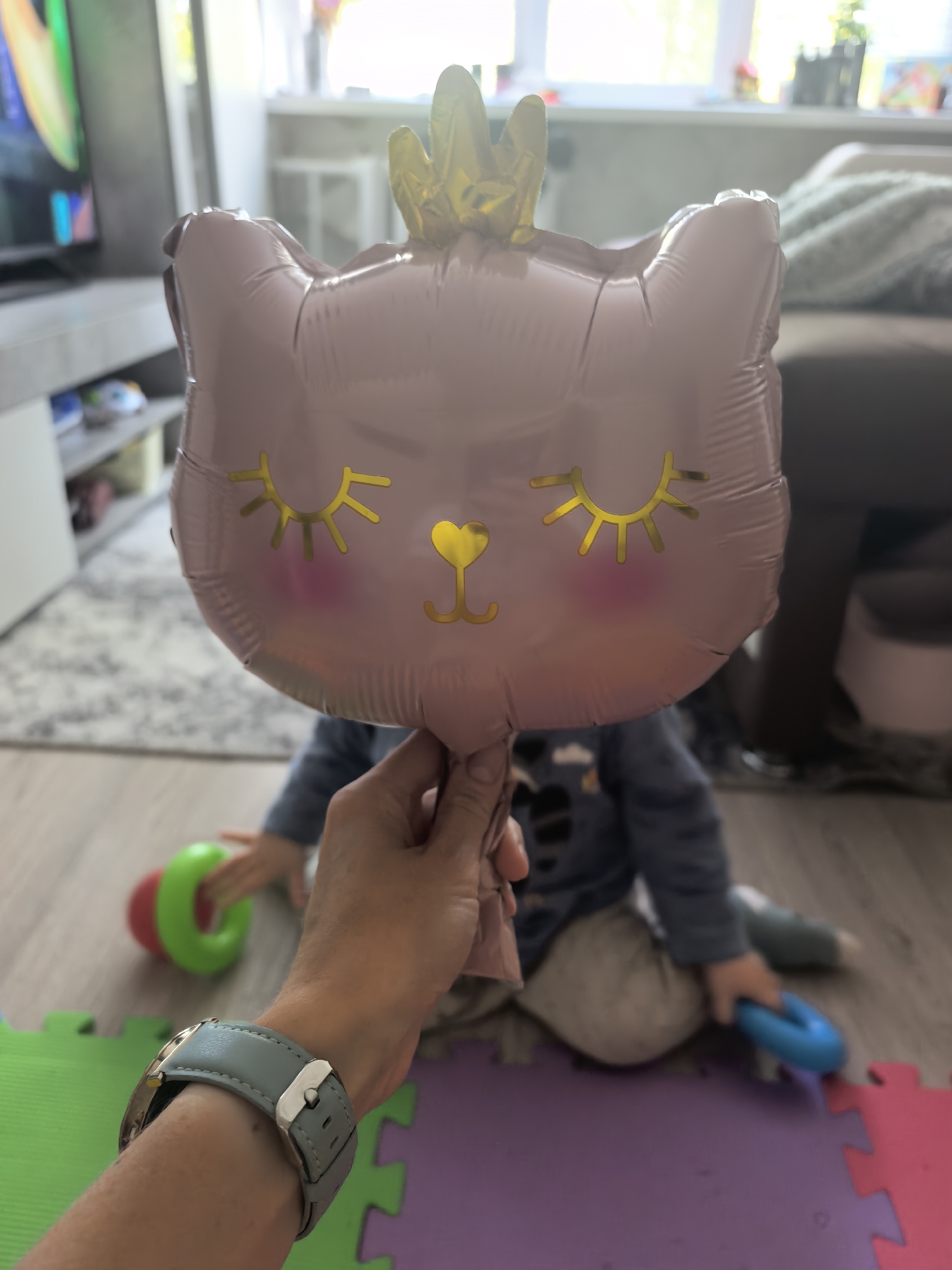 A hand holds a pink, cat-shaped balloon with a golden crown. The balloon features closed, golden eyes and a small golden heart for a nose. A child is seated on the floor behind the balloon, on interlocking foam play mats of green, purple, and red. The child wears a long-sleeved blue shirt and light-colored pants, and is playing with a blue ring. A green and red toy is visible on the left. The hand wearing a watch with a light blue band is holding the balloon. In the background, a television sits on a cabinet, with a blurred couch and window visible further back. The location appears to be indoors. The city of Sterlitamak, Russia is not discernible from the image.