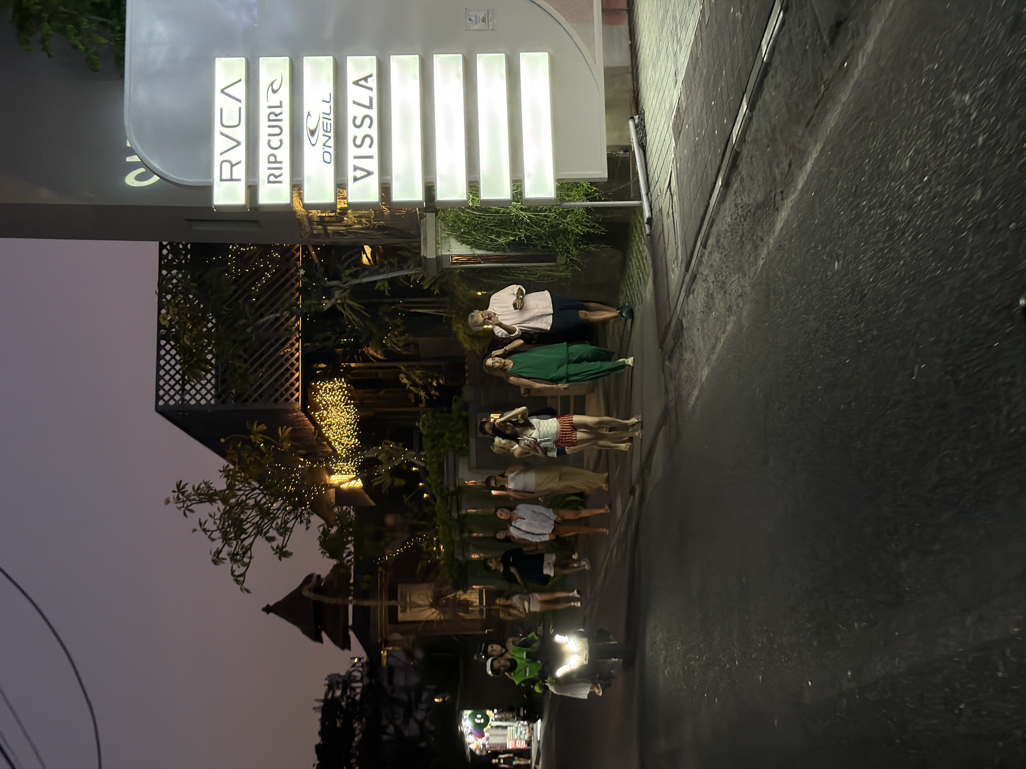 In North Kuta, Indonesia, at dusk, a group of people walks along a dimly lit street next to a building adorned with illuminated signs. The building features several illuminated rectangular signs displaying brand names such as RVCA, RIPCURL, O'NEILL, and VISSLA. A scooter with a rider and a passenger in a green vest passes by on the street. The building also has decorative string lights draped around its structure. The atmosphere is reminiscent of a commercial area in a tropical location at the end of the day.