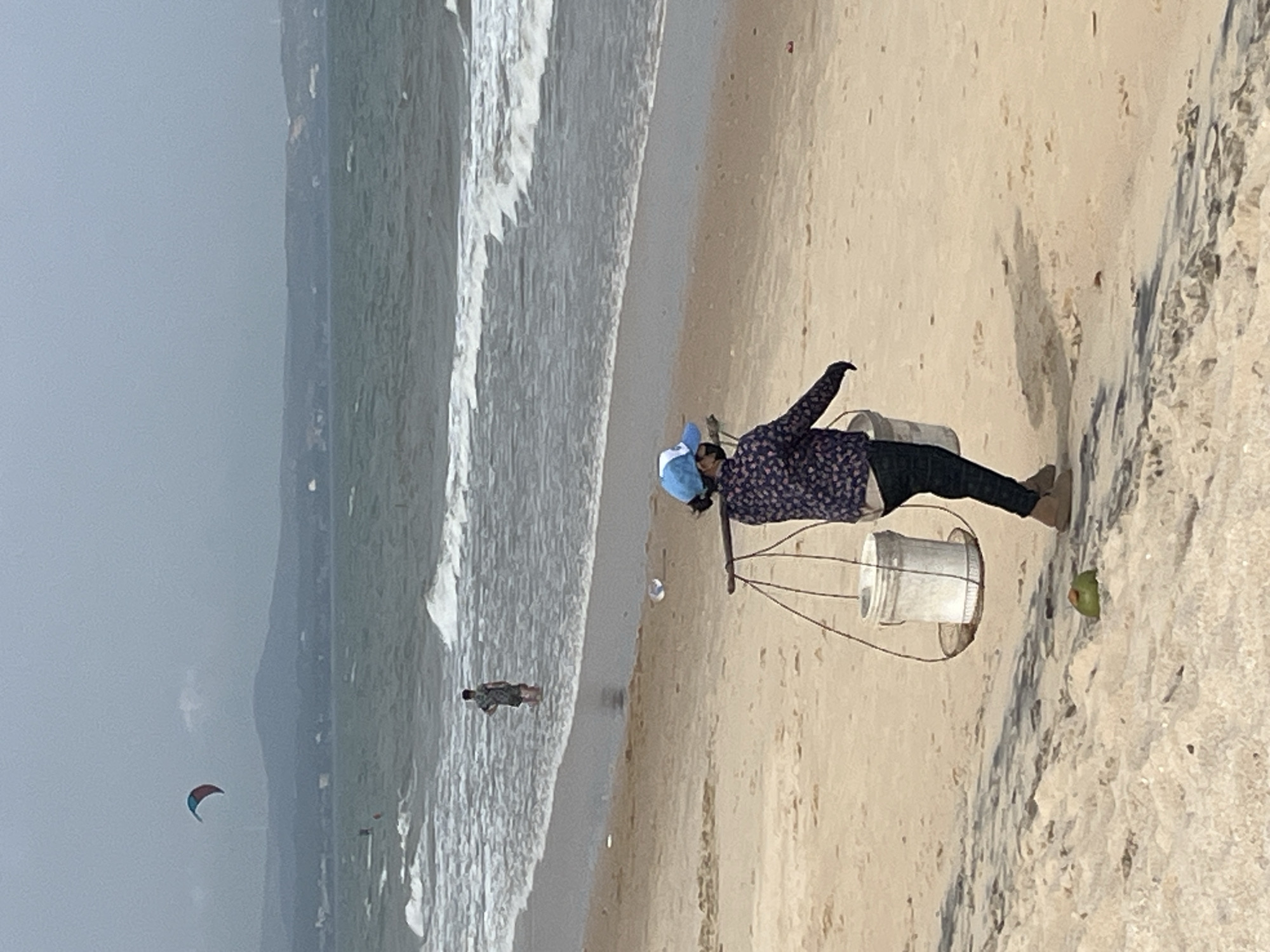 The image depicts a beach scene in Mũi Né, Vietnam. A person wearing a blue cap and a patterned shirt walks on the sand, carrying a yoke balanced on their shoulders with two white buckets hanging from it. In the background, the ocean waves crash onto the shore, and a person stands in the water. Further out, a kite is visible in the sky above a mountainous landscape. The beach is sandy and appears somewhat overcast.