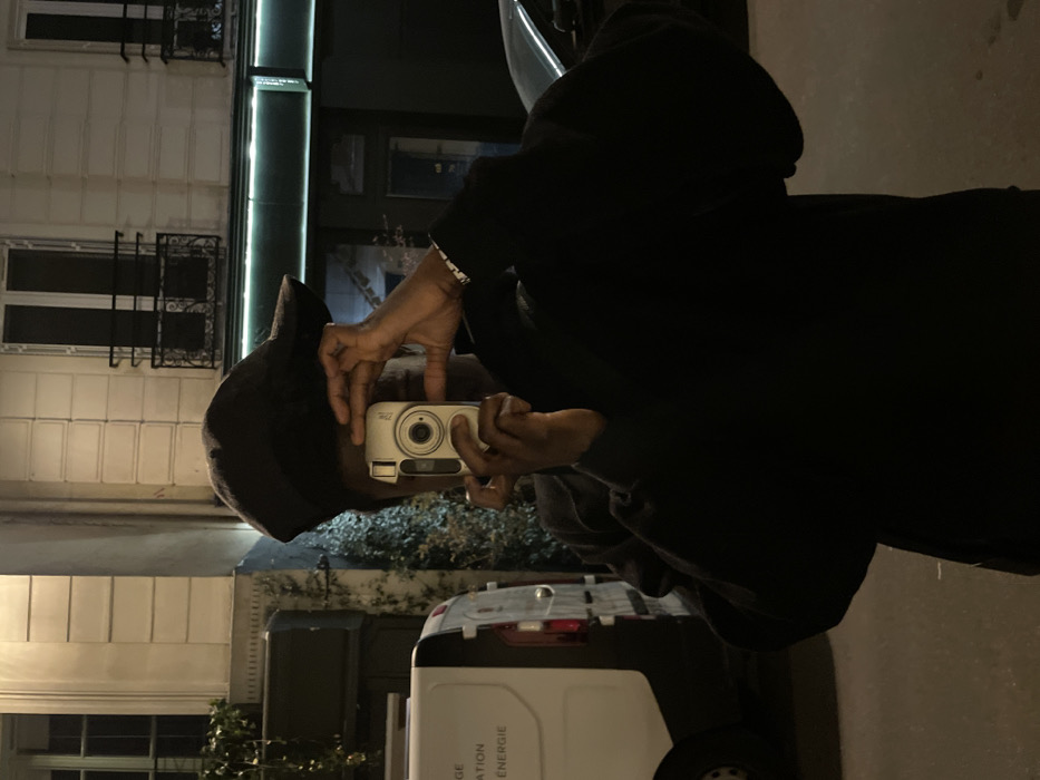 The image features an individual with dark skin, positioned on an urban street at night in Paris, France. The person's face is entirely obscured by a silver-colored compact digital camera held directly in front of them, with both hands operating the device. They wear a dark baseball cap and a dark long-sleeved top or jacket, revealing a patterned white cuff on the right wrist.
To the left of the individual, the rear portion of a white panel van is visible, displaying partial French text, specifically "AGE", "ATION", and "ÉNERGIE". Green foliage grows on a building wall behind the van.
The background is dominated by a light-colored Parisian building facade. Two upper-story windows with dark frames and ornate black wrought-iron Juliet balconies are prominent. Below these, a dark green ground-level facade or shopfront features a horizontal green neon light strip. A dark vehicle is partially discernible on the far right. The ground is a dark asphalt street surface.