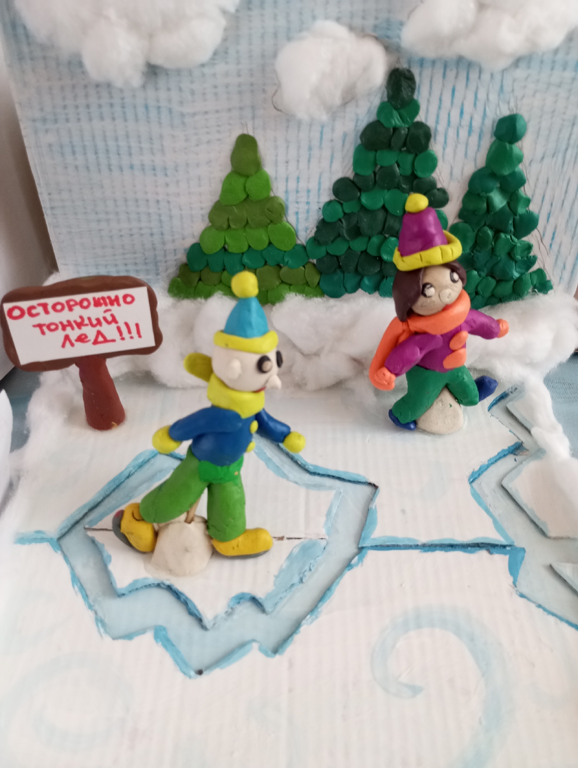 This is a vibrant, handmade diorama or craft project depicting a winter scene, likely focusing on ice skating. The scene features two humanoid figures, possibly children, crafted from colorful clay or plasticine, dressed in warm winter attire.
The figure on the left wears a blue and yellow hat, a yellow scarf, a blue jacket, green pants, and yellow shoes or skates. This character is positioned over a section of ice that appears to be cracking and breaking, with a piece of light-colored material (possibly clay or cotton) beneath one foot suggesting instability or a fall. The second figure, on the right, wears a purple and yellow hat, an orange scarf, a purple jacket, green pants, and blue shoes/skates, standing on a more stable section of the icy surface. Both figures have simple, round faces with dot eyes.
In the background, three dark green fir trees, also made from layered clay/plasticine pieces, stand on snowy ground. The sky is a light blue with painted vertical lines, and fluffy white cotton clouds are affixed to it. The foreground consists of a white surface representing snow-covered ice, with painted blue outlines indicating ice formations and deep cracks. White cotton is also used to create snowdrifts around the trees and along the edges of the scene, enhancing the winter atmosphere.
A notable detail is a small, brown signpost on the left side. The sign bears a warning in red capital letters: "ОСТОРОЖНО ТОНКИЙ ЛЁД!!!", which translates to "CAUTION THIN ICE!!!". This sign provides a narrative element, highlighting the potential danger of the scene, particularly for the figure whose ice is breaking. The craft uses materials like clay/plasticine, cotton, and painted cardboard for the base and background. The overall impression is a charming yet cautionary winter artwork, set in a snowy, outdoor environment, possibly a frozen pond or lake. The location context is Izhevsk, Russia.