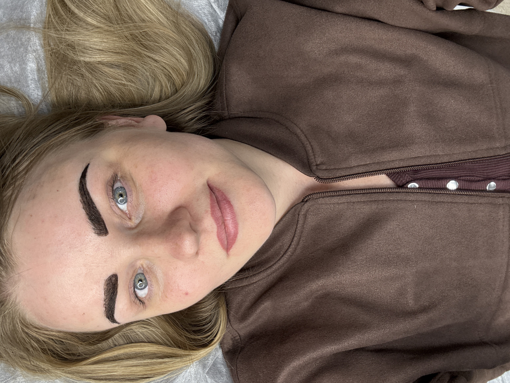 A woman lies on a light grey surface, presumably a treatment table, with her head positioned at the top left of the frame. She has blonde hair, fair skin, and blue eyes. Her eyebrows appear to be recently tattooed or cosmetically enhanced, exhibiting a dark, defined shape. Her lips also appear to have been cosmetically enhanced. She is wearing a brown zippered sweatshirt, revealing a darker brown undershirt with silver buttons. The location is not specified and cannot be determined from the image alone.