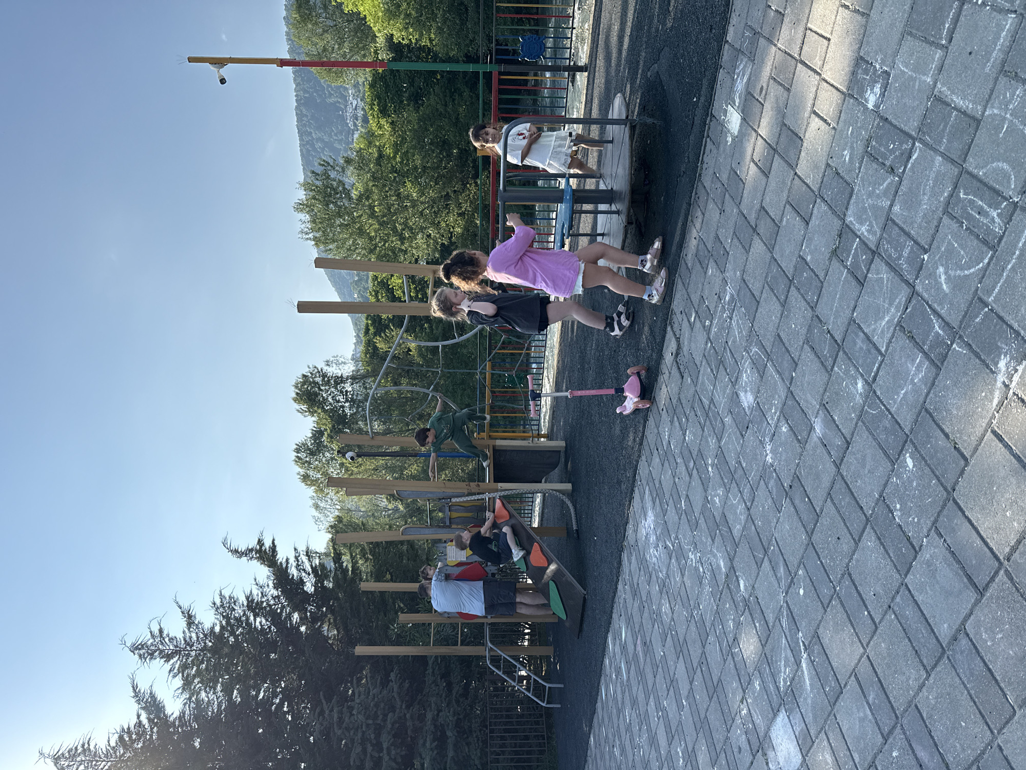 The image depicts a children's playground situated in Sochi, Russia, on a clear, sunny day. The playground features a multi-level structure with slides, climbing elements, and a merry-go-round. Several children are actively engaged in play: a boy in a green outfit is jumping from a platform, another boy in a black t-shirt is climbing a slide, and three girls are interacting near the merry-go-round. An adult male in a blue t-shirt and shorts is also present, supervising near the climbing structure. A pink scooter is parked on the side. The playground is surrounded by lush greenery and a body of water, possibly a river, is visible in the background. The ground is paved with interlocking gray bricks, with some chalk drawings scattered across them. A security camera is mounted on a pole in the background.