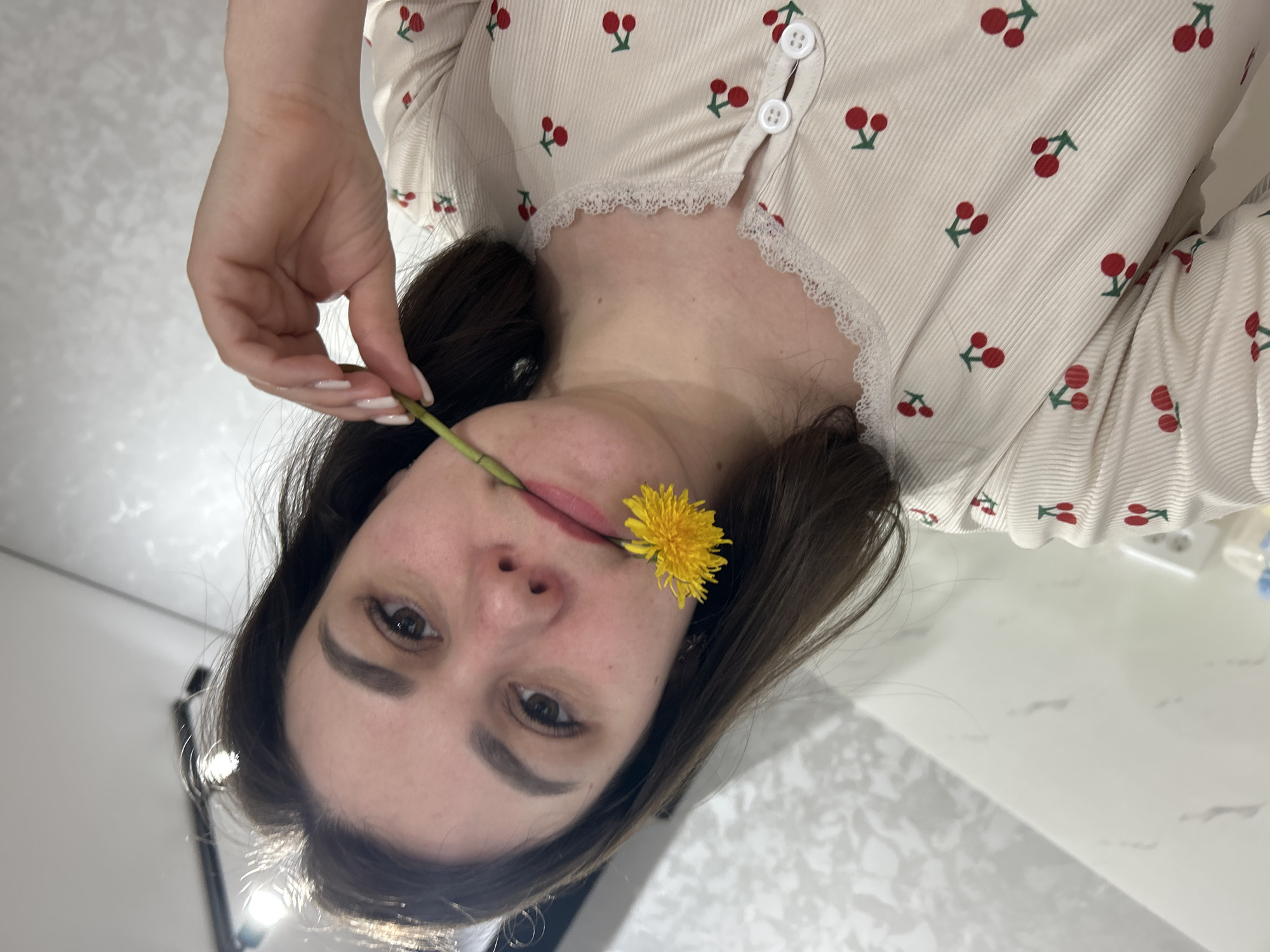 A woman is holding a dandelion in her mouth. She is wearing a cream-colored, cherry-patterned shirt with lace detailing around the collar. Her left hand holds the stem of the dandelion. The location is likely an indoor setting in the city of Chaikovsky, Russia.