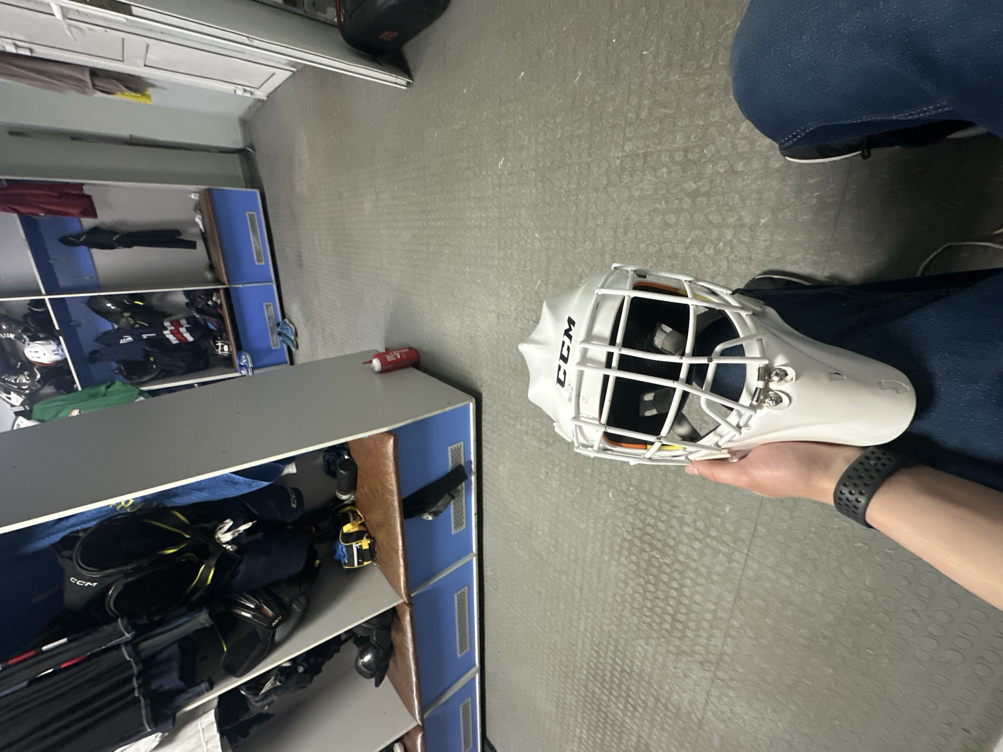 The image shows an indoor space with lockers lining the walls. The floor is gray with a repeating circular pattern. A person wearing blue jeans is partially visible in the upper right. A white CCM hockey helmet is being held in the person's right hand, which also has a black smart watch on the wrist. A tube of some sort is on top of the locker. The lockers are mostly gray with blue trim and brown interiors. Inside the lockers are various pieces of equipment. The location of this image is Unknown, Unknown.