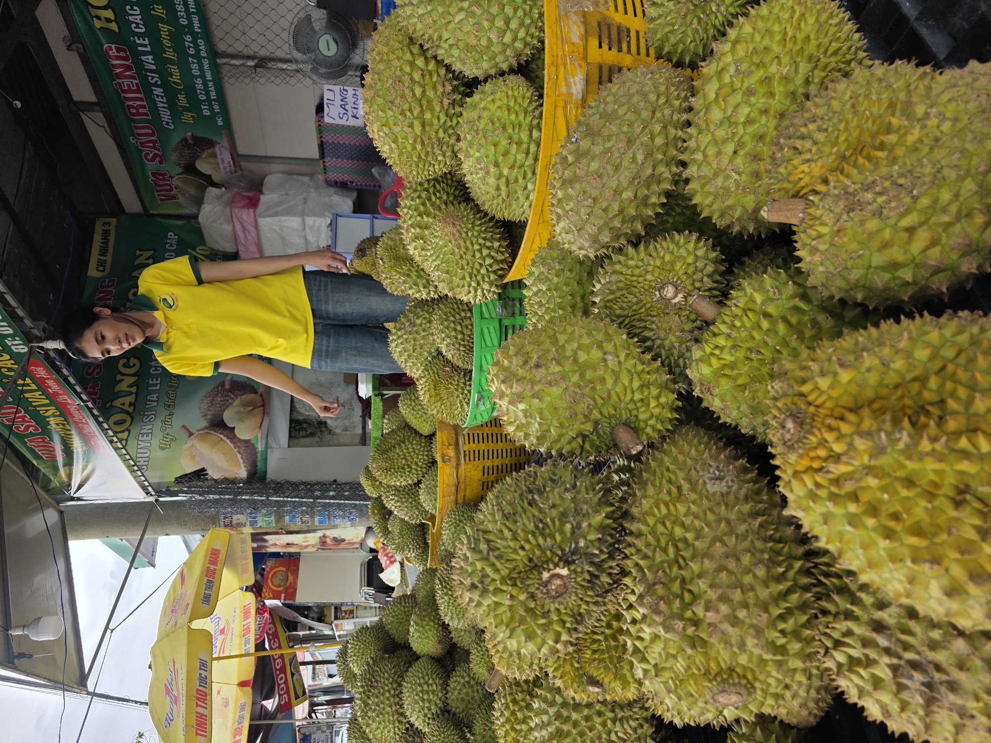 Here is a matter-of-fact description of the image:
The image shows a street-side durian stand in Phu Thuy Ward, Vietnam. Large piles of durian fruit, some in yellow plastic crates, dominate the right side of the image. To the left, a woman in a yellow shirt and jeans stands near the durian display. Behind her are vendor signs and umbrellas. A banner in Vietnamese promoting "Vua Sau Rieng" (King of Durian) is visible. The text on the banner indicates business hours and contact information, including the address "107 Tran Hung Dao - Phu Thuy." Several commercial posters and advertisements are also in view. The overall setting seems to be a bustling commercial area.
