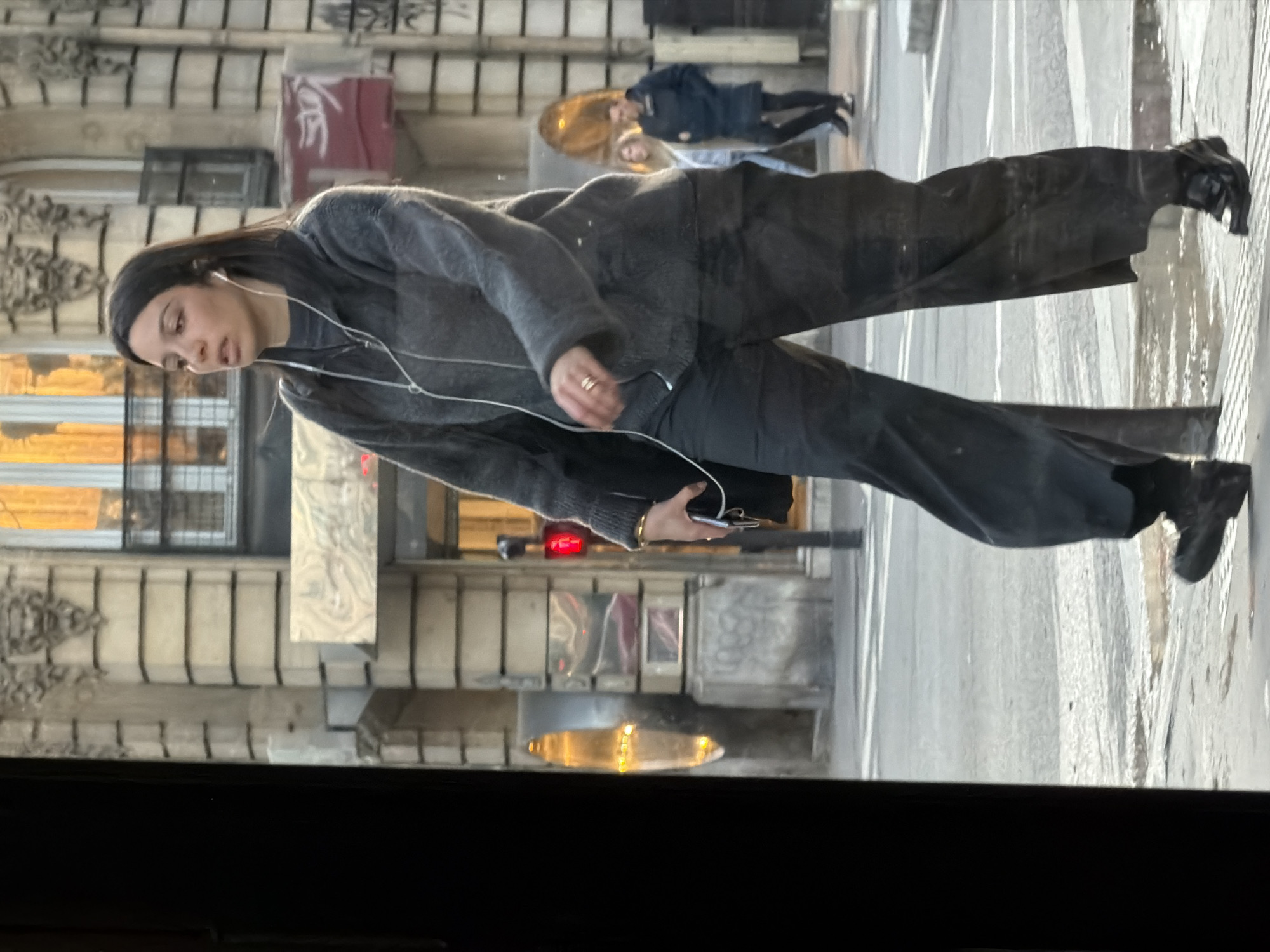 A young adult female is captured mid-stride, walking from right to left across a street or pavement in Paris, France. She has long dark hair, light skin, and is looking slightly upwards and to her left with her mouth slightly open. She wears a dark grey, zippered, textured sweater over a darker top, loose-fitting dark trousers, and dark, polished shoes. White wired in-ear headphones are visible, with the wire extending down her front. A gold-colored ring is on her right ring finger. In her left hand, she carries a black shoulder bag and a dark-colored smartphone.
The immediate ground beneath her feet shows wet patches on the pavement and street, suggesting recent moisture. Faint white crosswalk markings are visible. In the background, a multi-story, light-colored stone building, characteristic of Haussmannian architecture, features horizontal banding and decorative elements, including a sculpted lion's head above a window. One window reflects a strong, warm orange light. To the main subject's right, a pedestrian traffic signal displays a red "stop" figure. To her left, a tall, yellow-orange street light or decorative lamp is visible. Further in the background, to the right, two additional individuals are visible: one person in a dark coat walking away from the camera, and another person with lighter hair in a light top, partially obscured. Indistinct red signage with white markings is visible behind the main subject's head. The scene is partially framed on the left by a dark, out-of-focus vertical element, likely a window frame or doorway.