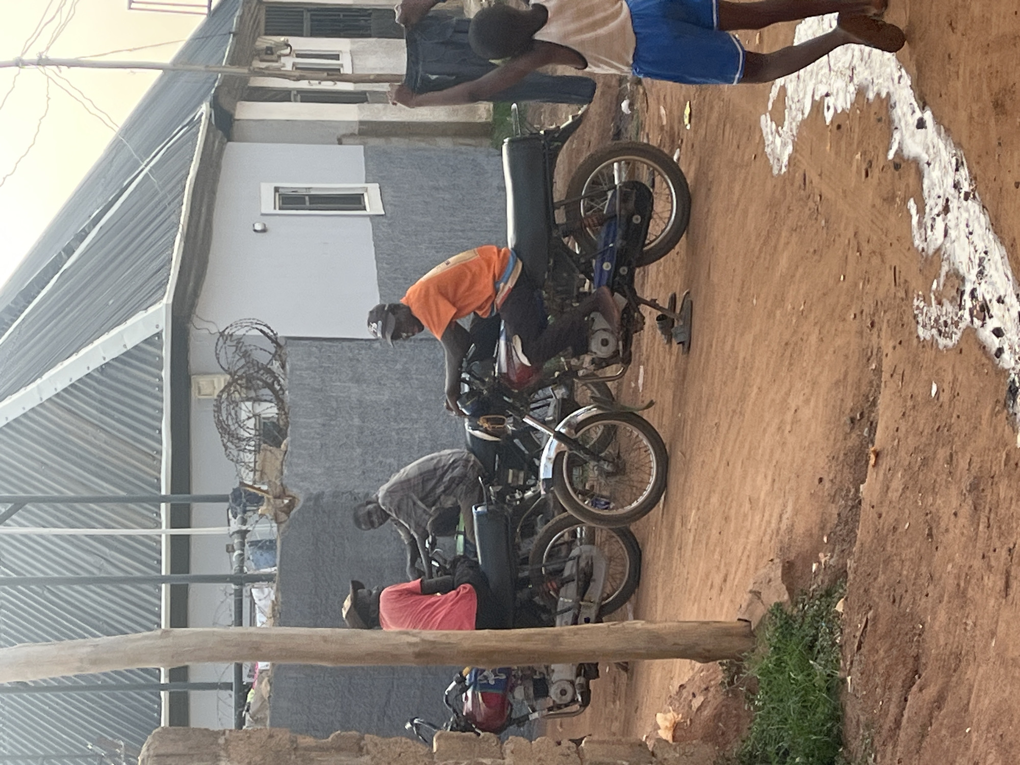 The image shows a scene in what appears to be a residential or commercial area in Abuja, Nigeria. The environment consists of a dirt road and buildings, one of which has a corrugated metal roof and a partially visible upper story with windows. Barbwire is visible near the buildings. There are several motorcycles parked along the road. At least three men are visible near the motorcycles, wearing casual clothing. One man is wearing an orange shirt, another a plaid shirt, and the third a red shirt. At the top of the image, two more people are present, one wearing blue shorts. Their interaction is not clear, but it appears to be a street scene. White debris or paint is visible on the ground on the far right of the image. A piece of lumber lies in the foreground.
